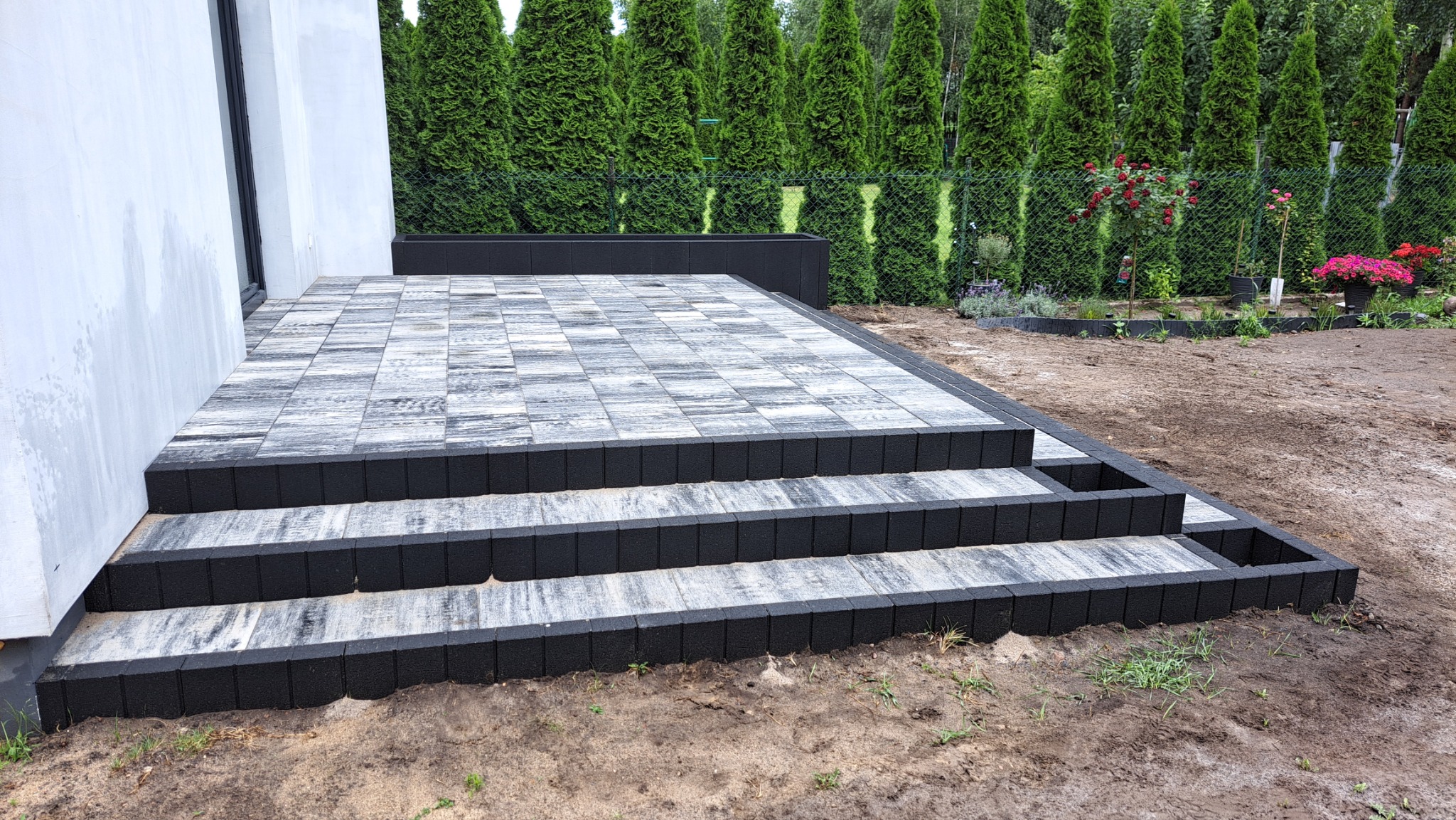 Schody i taras z szarej kostki brukowej z czarnym obramowaniem, przylegające do białego budynku, z widokiem na zielony ogród z tujami w tle.