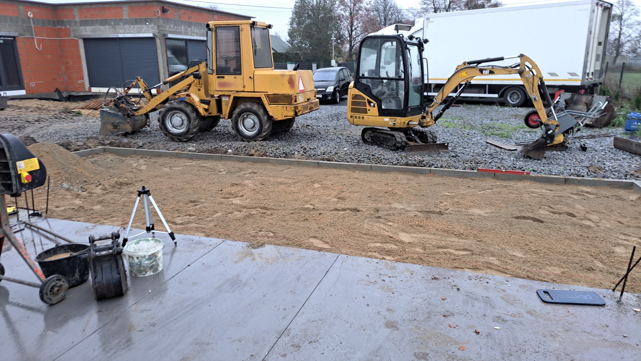 Przygotowanie podłoża pod kostkę brukową: żółty traktor i mini koparka CAT na budowie, widoczne obrzeża, piasek, gruz, mieszalnik do betonu, poziomica i inne narzędzia.