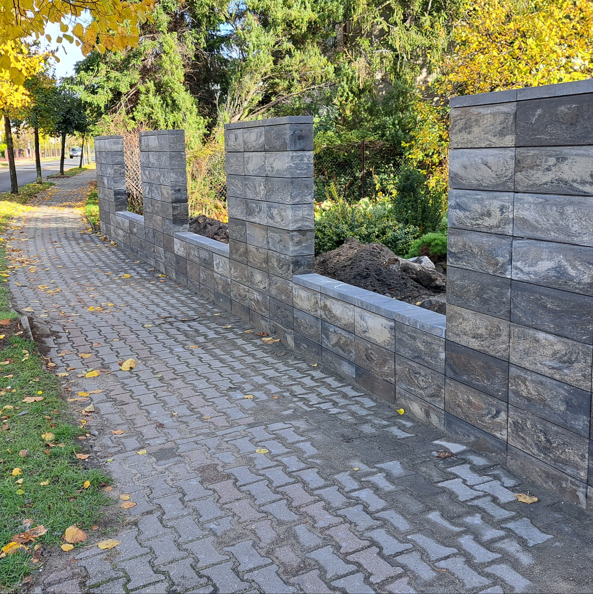 Nowo wzniesiony, modułowy mur ogrodzeniowy z szarej, melanżowej kostki, oddzielający posesję od chodnika wyłożonego kostką brukową, z jesiennymi liśćmi na powierzchni.