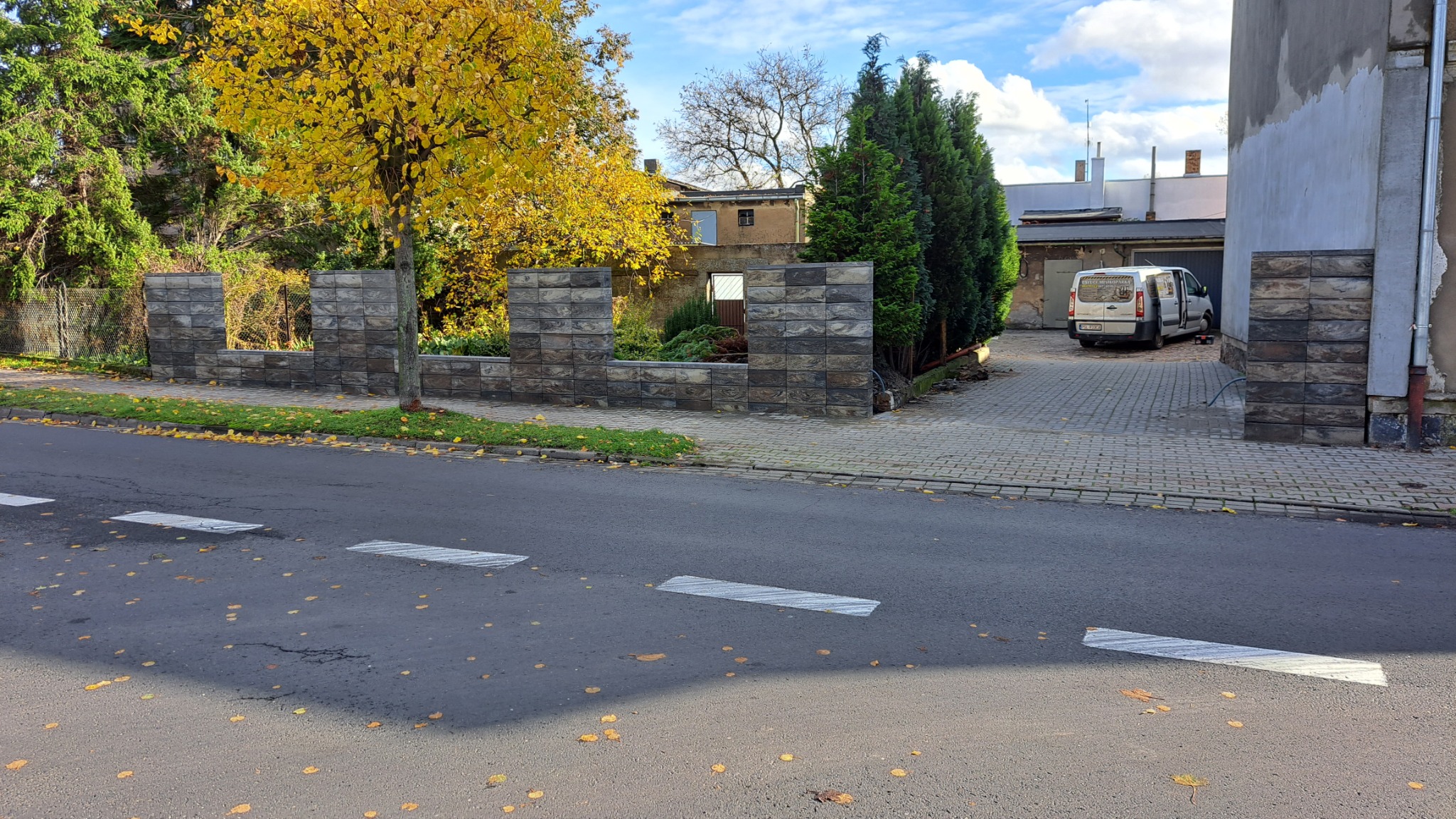 Nowoczesny, częściowo ukończony murek z szarej kostki łupanej, oddzielający posesję od ulicy, z widocznym samochodem dostawczym w tle na kostce brukowej.