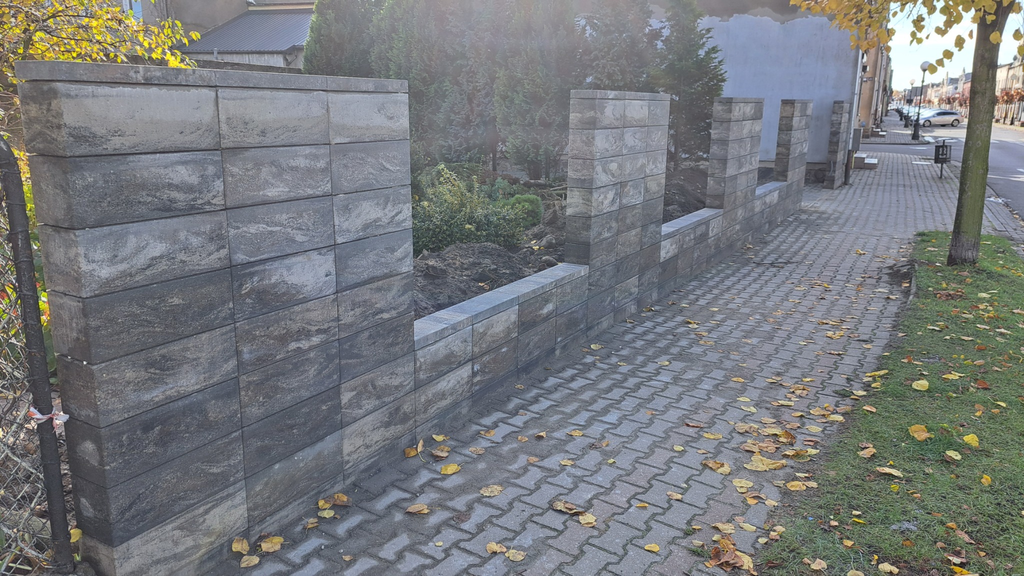 Nowoczesny, częściowo ukończony mur z szarej kostki łupanej, oddzielający posesję od chodnika wyłożonego kostką, z widoczną ziemią wewnątrz i drzewami w tle, w jesiennej scenerii z opadłymi liśćmi.
