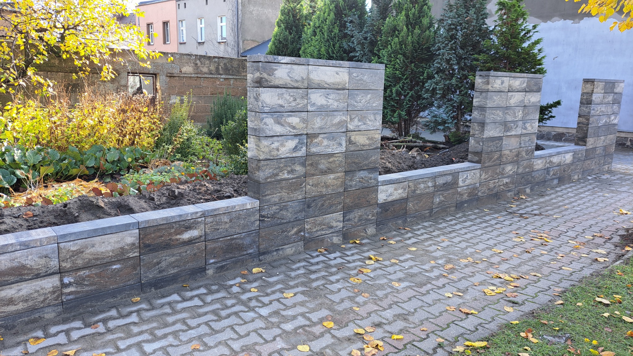 Nowo wybudowany murek z szarej kostki imitującej kamień, oddzielający ogród od chodnika z kostki, z widocznymi jesiennymi liśćmi na chodniku i zielenią w ogrodzie.