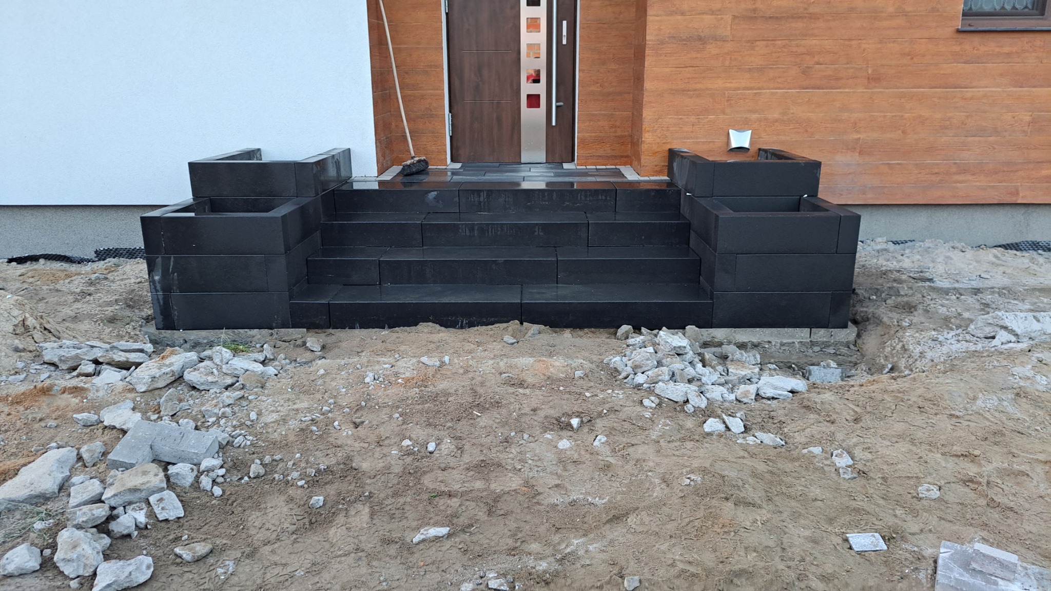 Nowe, czarne schody z kostki brukowej prowadzące do drzwi wejściowych domu, otoczone ziemią i gruzem przed zagospodarowaniem terenu.
