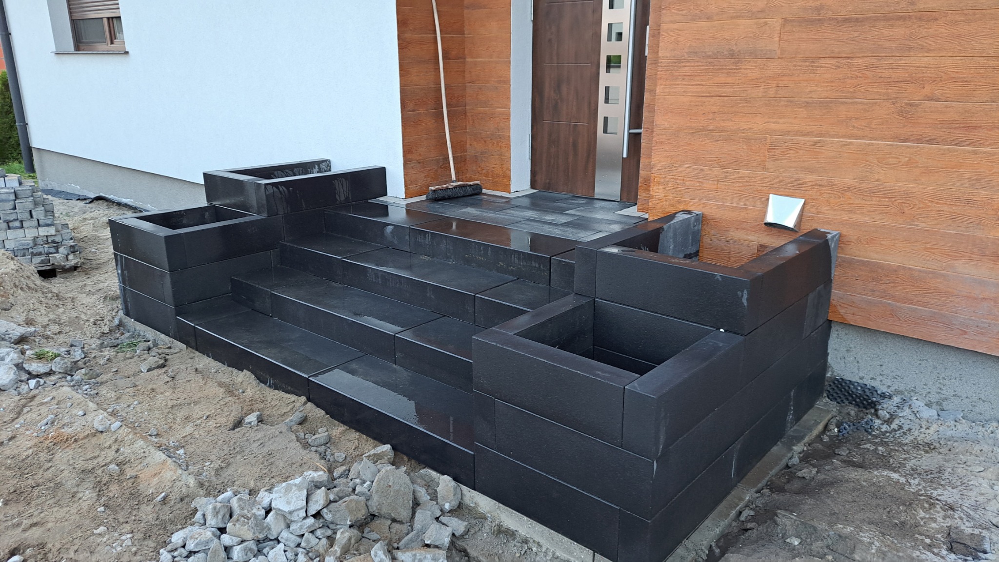 Nowoczesne, czarne schody wejściowe z kostki brukowej, częściowo ukończone, z widocznym stosem kostki obok i szczotką opartą o ścianę budynku.