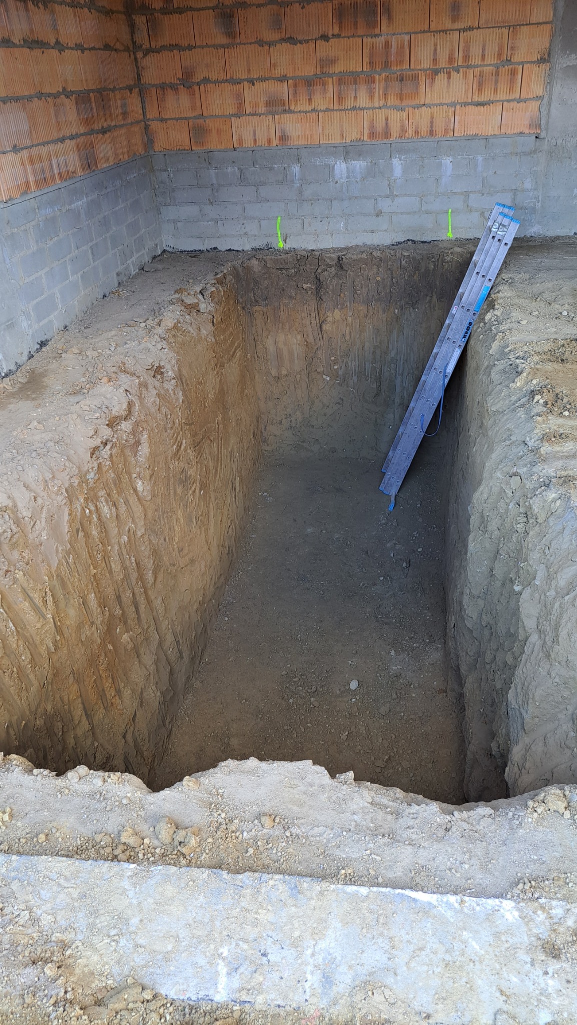 Głęboki wykop w ziemi z drabinką opartą o ścianę, widoczne bloczki betonowe i cegły na górze, zielone oznaczenia na ścianie wykopu.