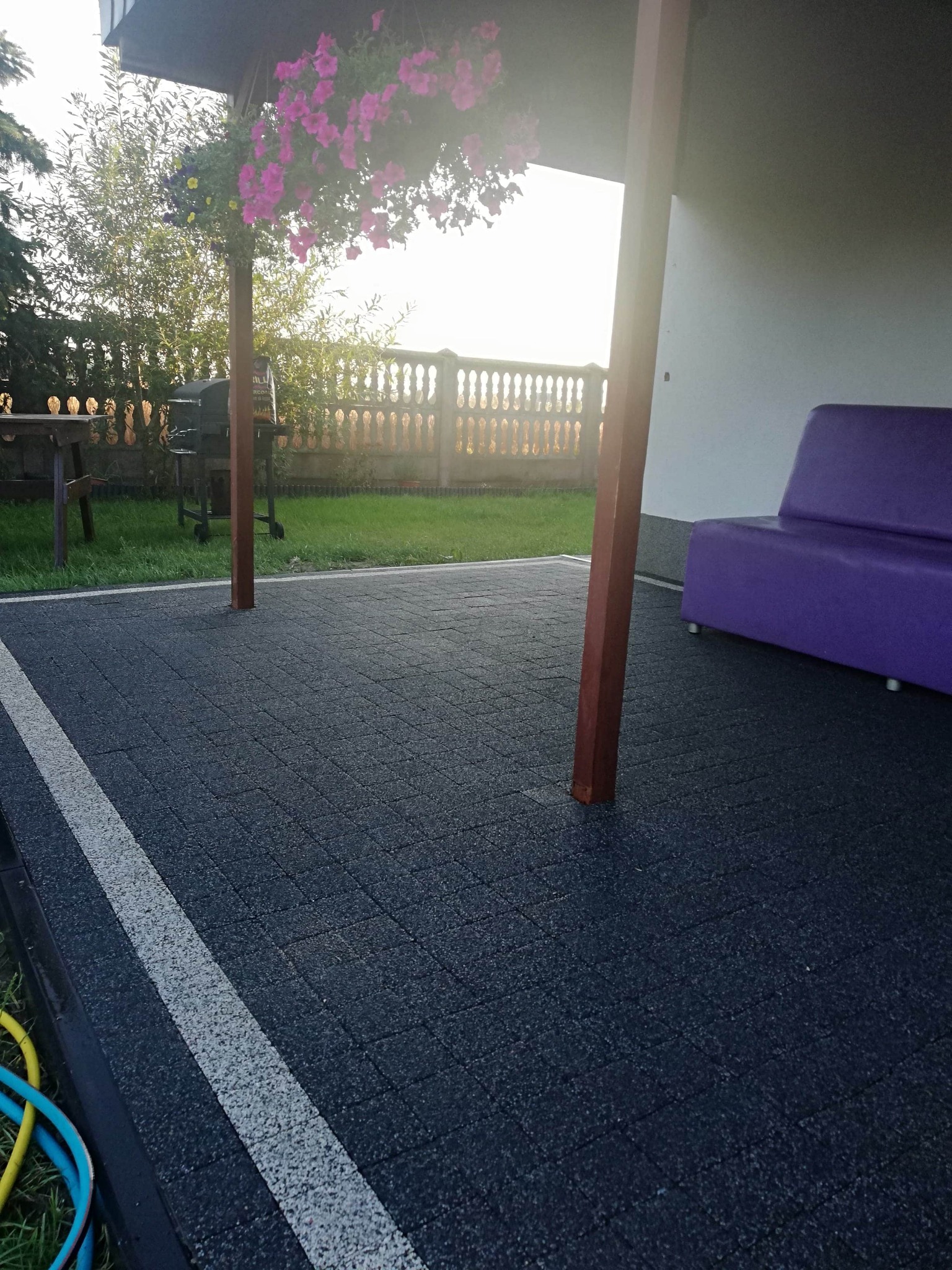 Ciemnoszara nawierzchnia z kostki brukowej z białym pasem oddzielającym, częściowo zadaszona weranda z fioletową kanapą w tle, kwiaty w wiszącej donicy.