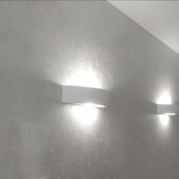 Empire Komfort Life - Ściana wykończona dekoracyjnym tynkiem w odcieniach szarości, oświetlona dwoma nowoczesnymi, białymi kinkietami.