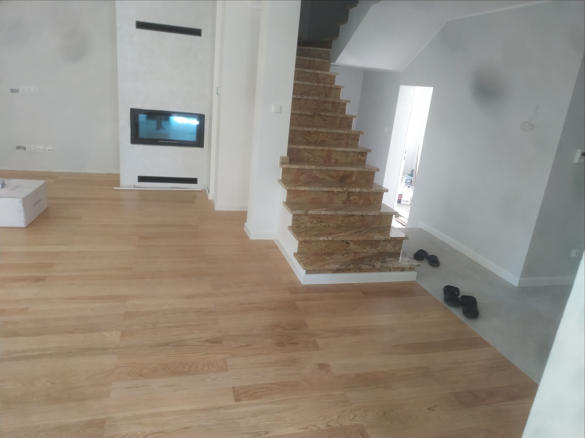 Jasne panele podłogowe i marmurowe schody w nowym domu w Warszawie, widok na kominek i świeżo pomalowane ściany.