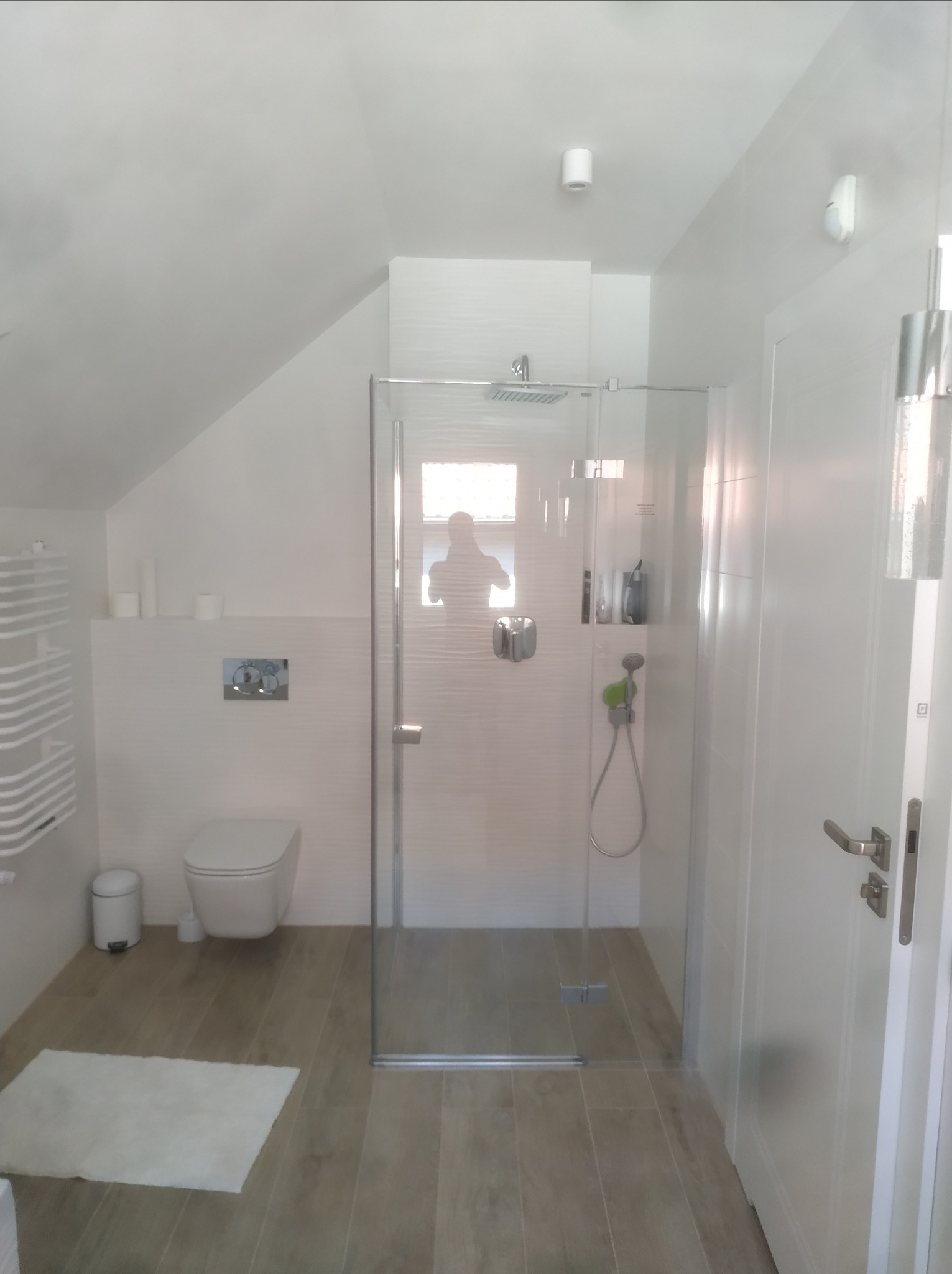 Nowoczesna, jasna łazienka na poddaszu z kabiną prysznicową typu walk-in, wiszącą toaletą, grzejnikiem drabinkowym i drewnopodobną podłogą.