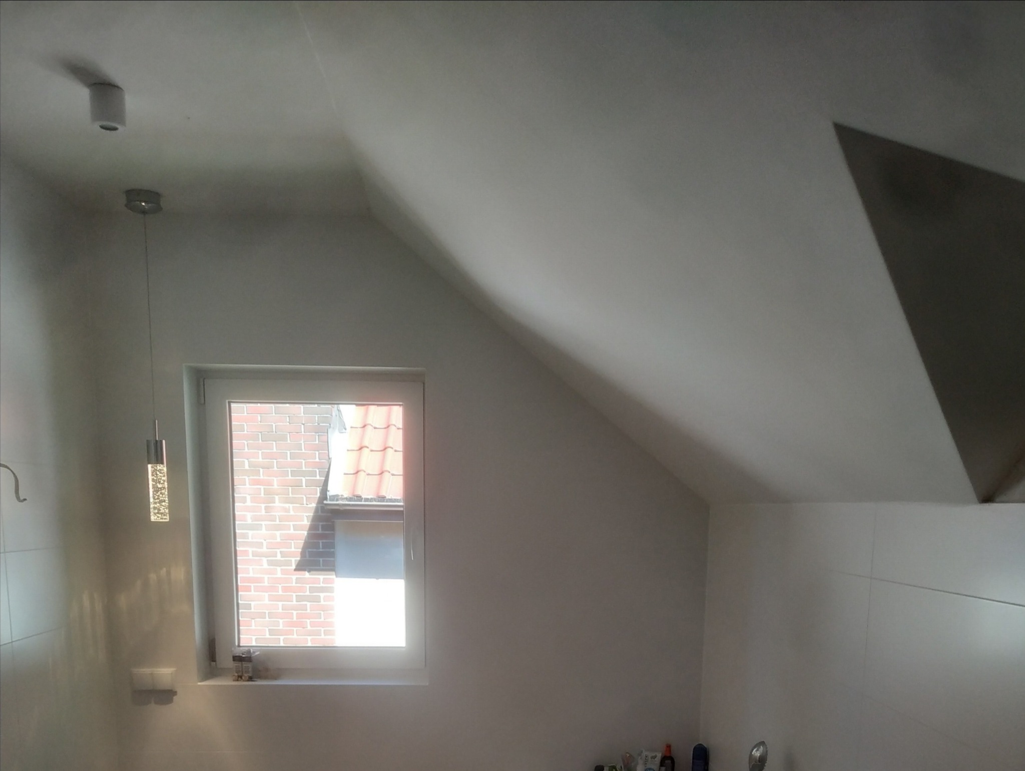 Pomieszczenie ze skośnym sufitem po świeżym odmalowaniu, widoczne okno z widokiem na ceglany budynek i dach z czerwonej dachówki, oraz nowoczesna lampa wisząca.