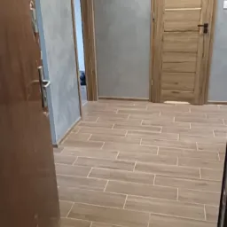 Korytarz po remoncie z drewnopodobną podłogą, szarą ścianą i dwiema drewnianymi drzwiami.