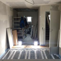 Pawlo ,glazurnictwo z pasją, - Instalacja ogrzewania podłogowego w surowym wnętrzu, widoczne rury grzewcze na podłodze, ściany w trakcie przygotowania do malowania, drabina i materiały budowlane w tle, dwie lampy budowlane...