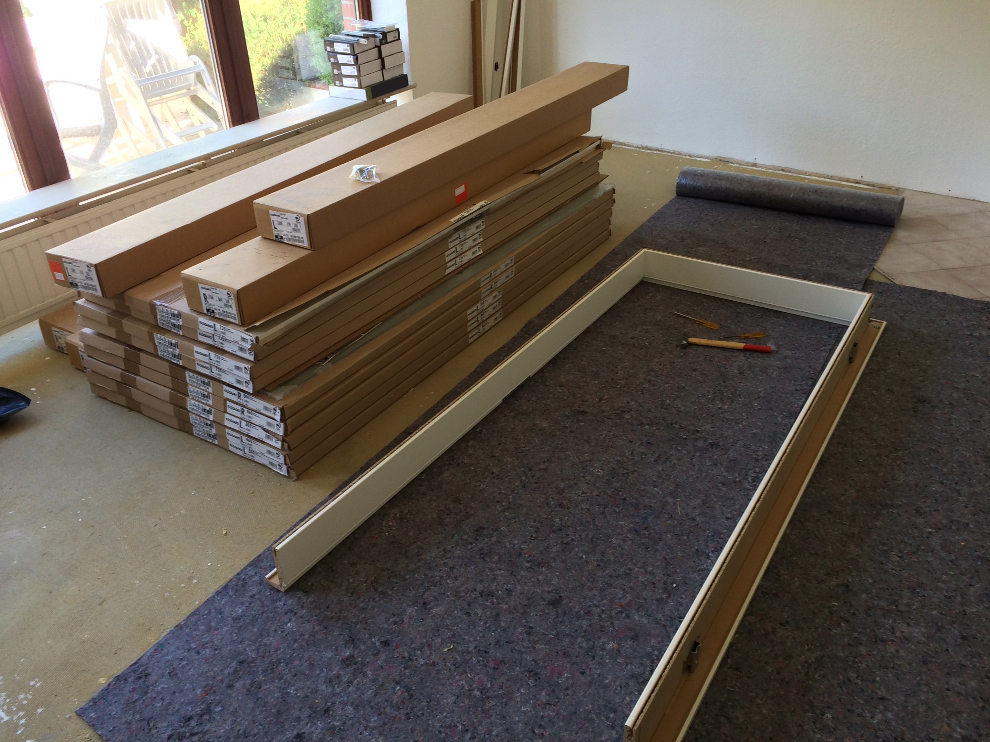 Przygotowanie do montażu podłogi: rozłożona mata ochronna, ułożona rama z paneli podłogowych, narzędzia i stos zapakowanych desek podłogowych przy oknie.