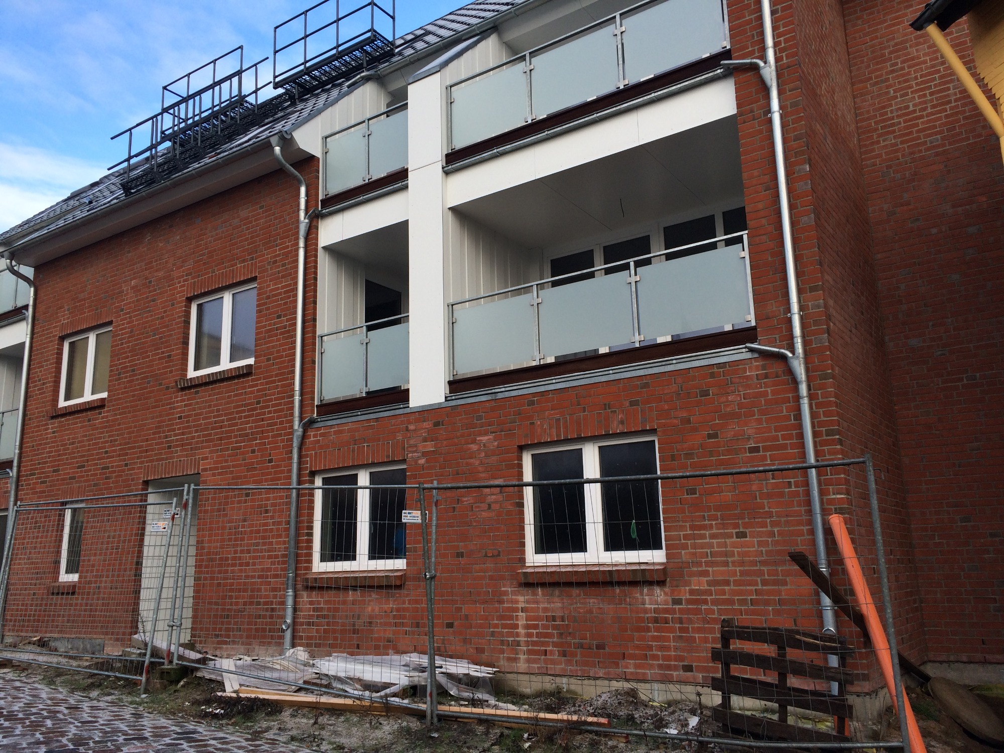 Nowy budynek mieszkalny z czerwonej cegły z balkonami z matowego szkła, zabezpieczony tymczasowym ogrodzeniem budowlanym.