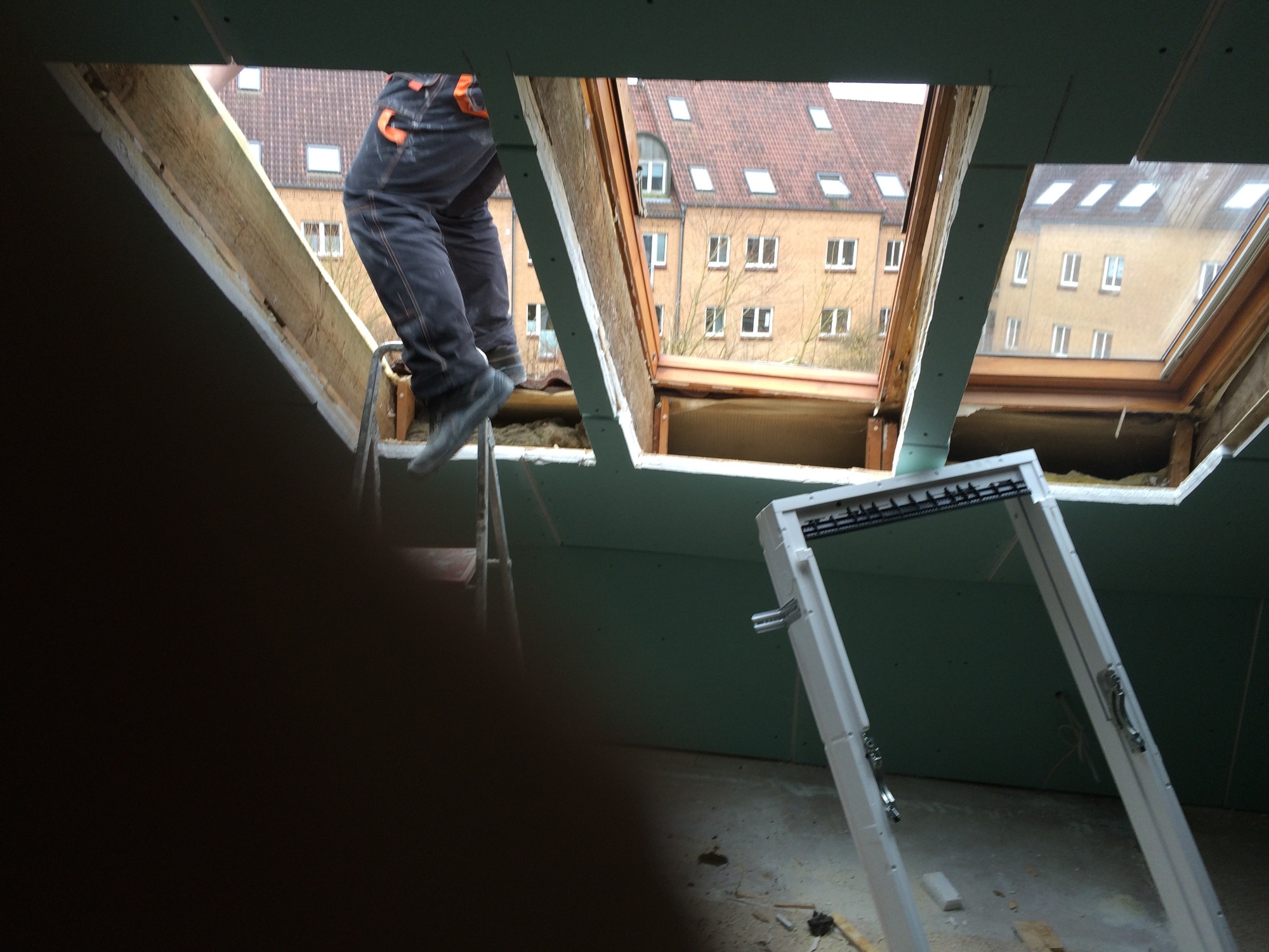 Montaż okna dachowego: monter na drabinie, widok na budynek z czerwonym dachem przez otwór okienny, rama okna oparta o ścianę z płyt gipsowo-kartonowych.