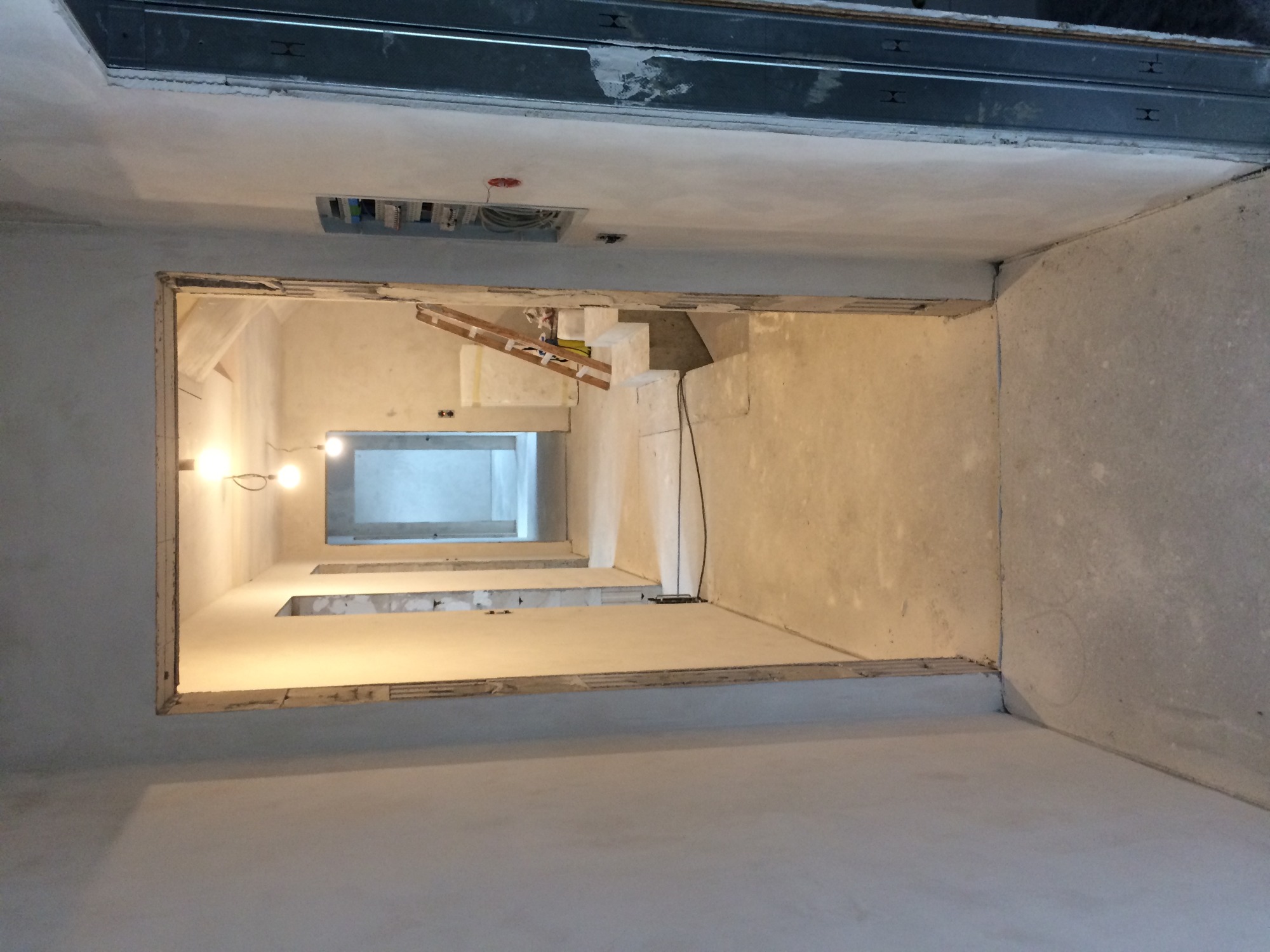 Długi korytarz w trakcie remontu z surowymi ścianami i widoczną instalacją elektryczną pod sufitem, oświetlony żarówkami.