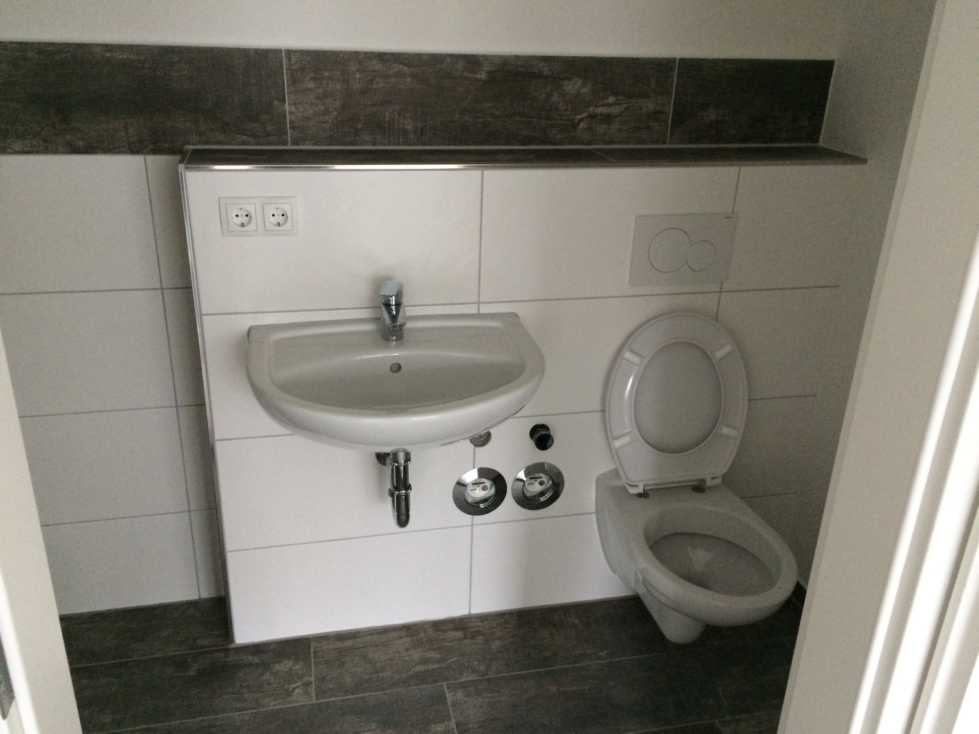 Nowoczesna łazienka z białymi płytkami, umywalką i toaletą podwieszaną, wykończona ciemnoszarą płytką dekoracyjną na górze. Widoczne dwa zawory pod umywalką z licznikami.