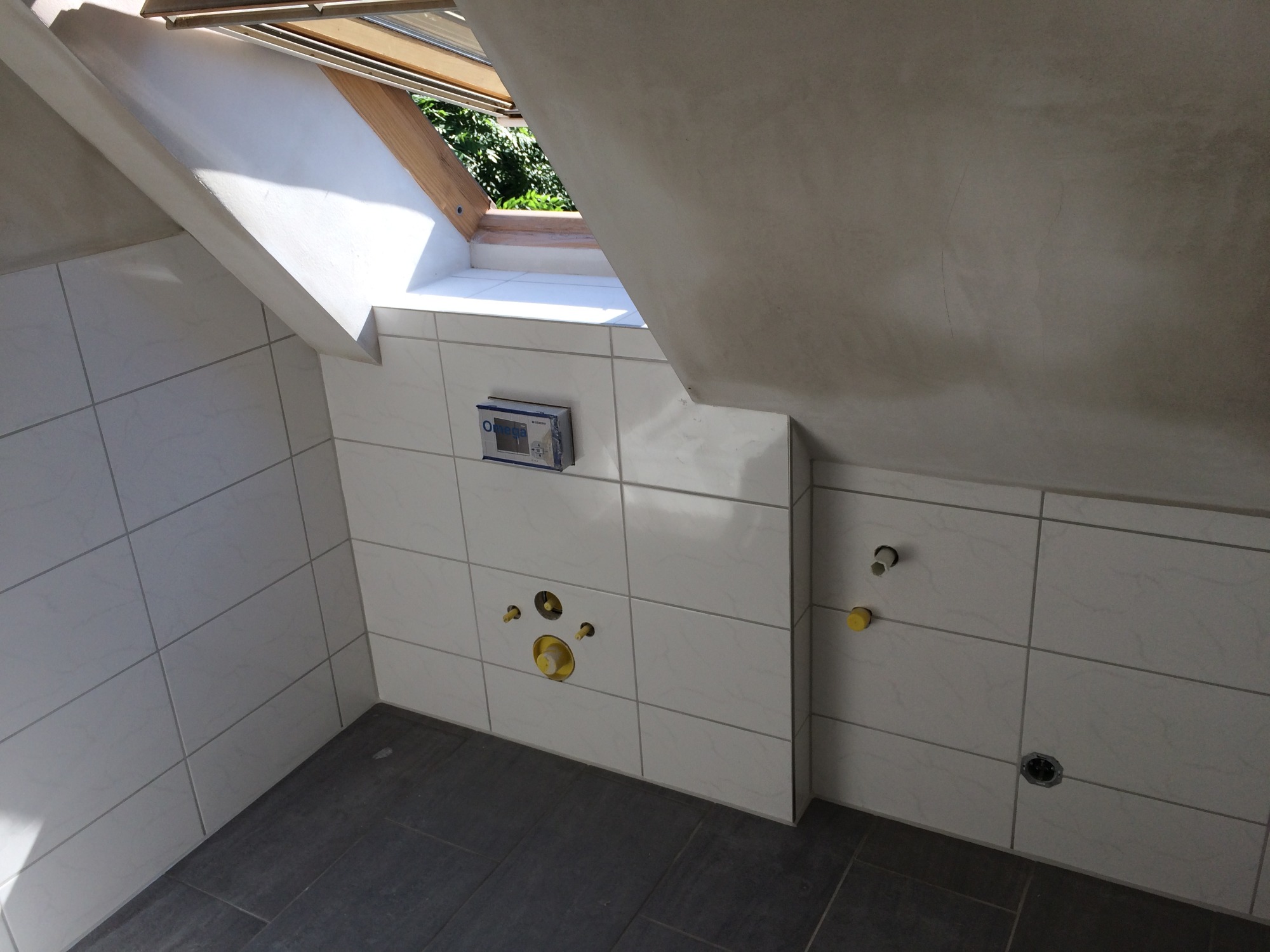 Wnętrze łazienki na poddaszu z oknem dachowym, wyłożonej białymi płytkami z szarą fugą i ciemną podłogą. Widoczne przygotowane instalacje wodno-kanalizacyjne oraz zamontowany panel sterujący.