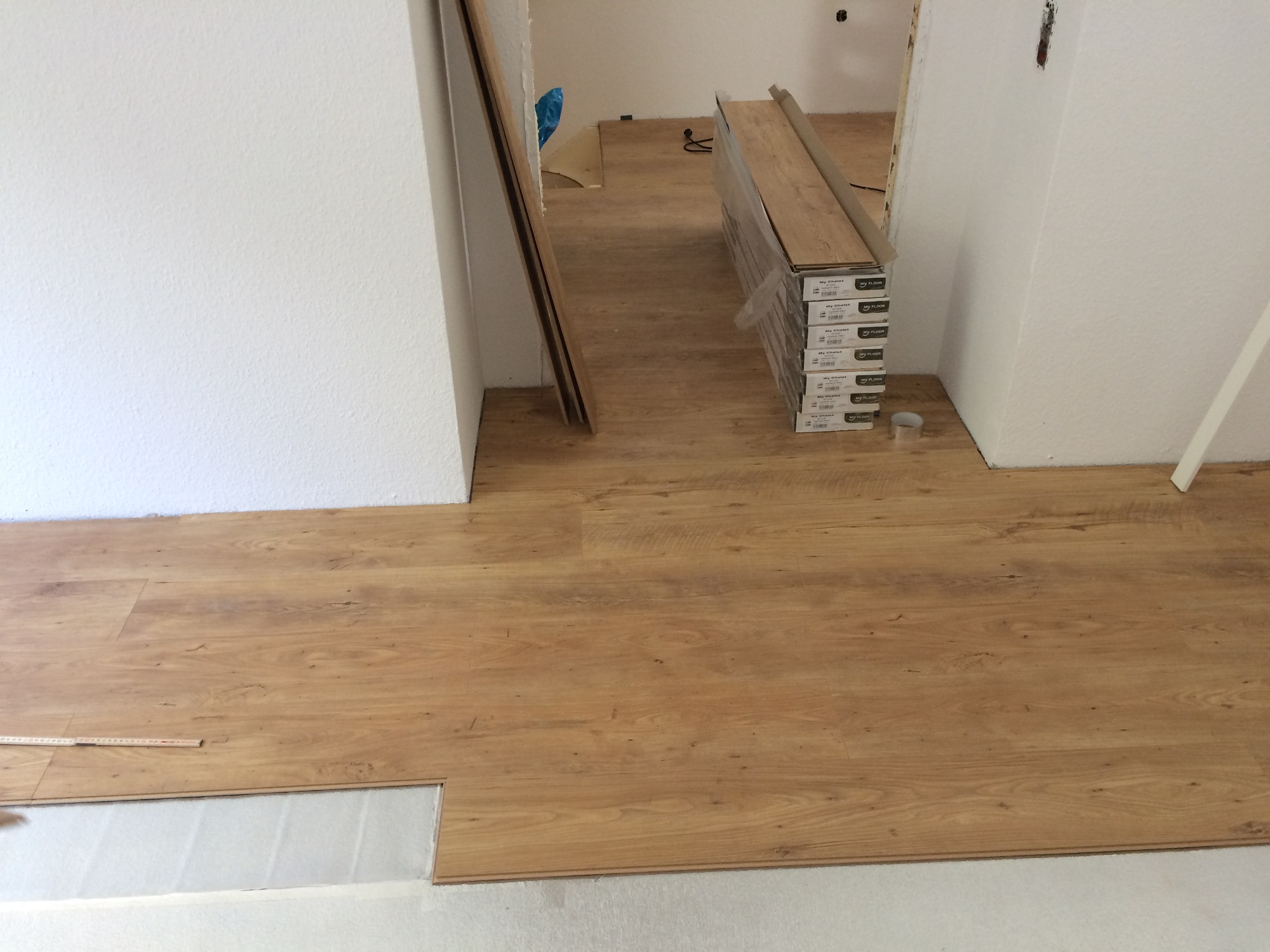 Układanie paneli podłogowych w jasnym odcieniu drewna, widoczne paczki paneli i fragment listwy przypodłogowej przy białej ścianie, w tle kolejne pomieszczenie.