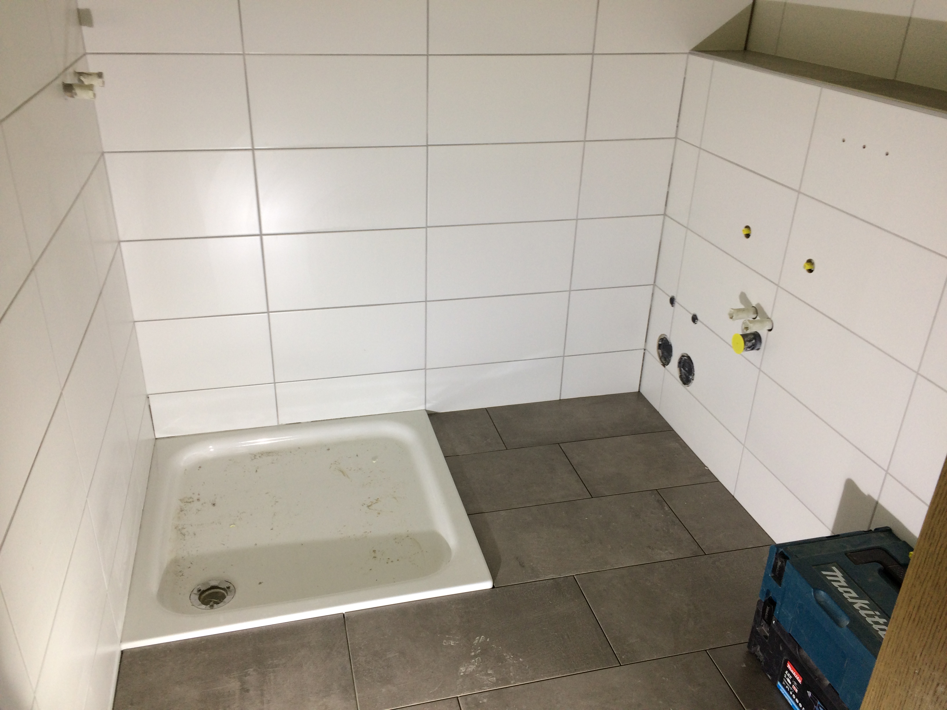 Przygotowana do położenia glazury kabina prysznicowa z odpływem, obok leżą szare płytki podłogowe, a na ścianach ułożone są białe płytki. Widoczne otwory na armaturę i skrzynka narzędziowa Makita...