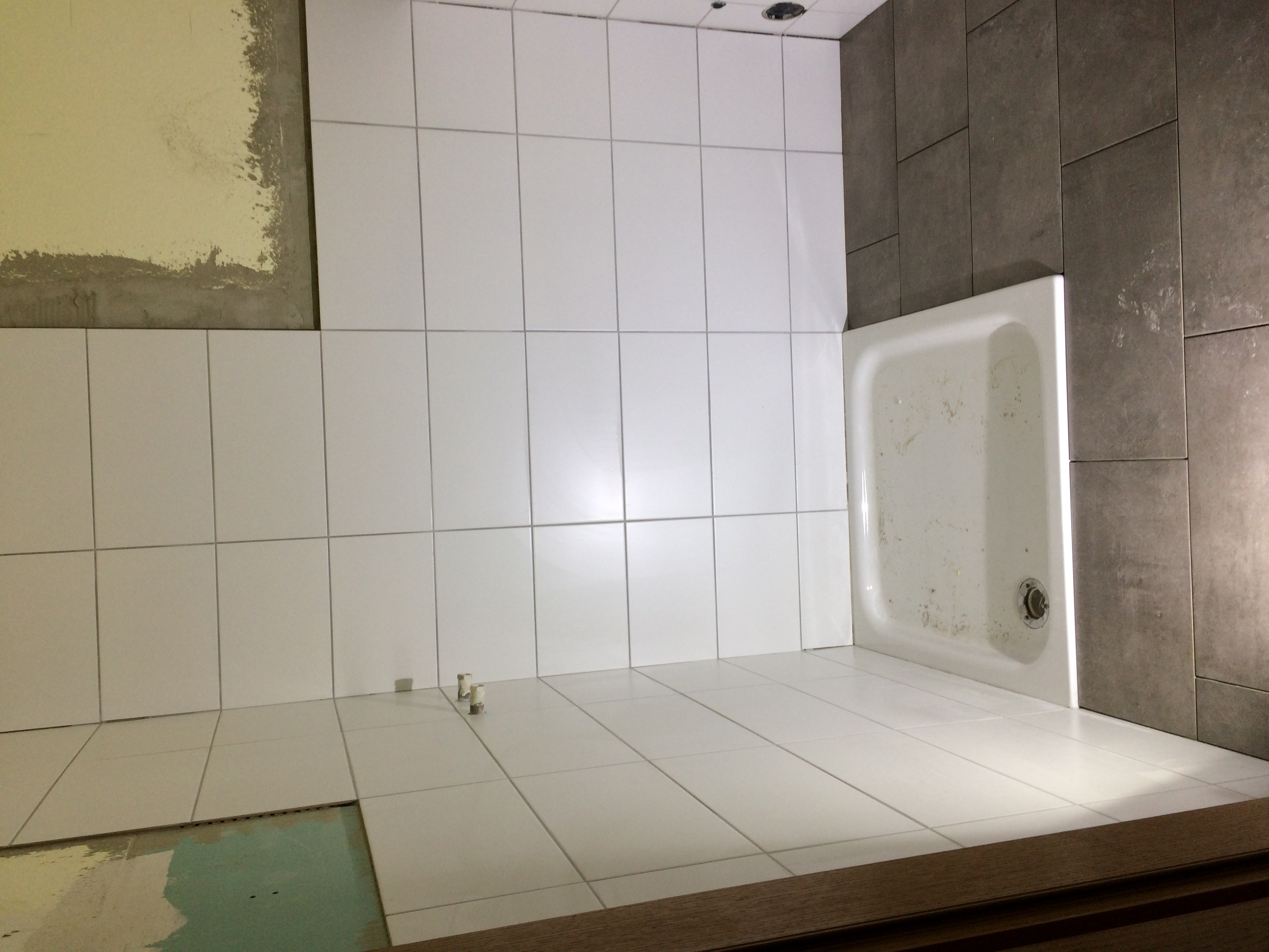 Niedokończona łazienka z białymi płytkami na ścianach i podłodze, widoczna biała brodzik, fragmenty ścian bez płytek i wystające rury.