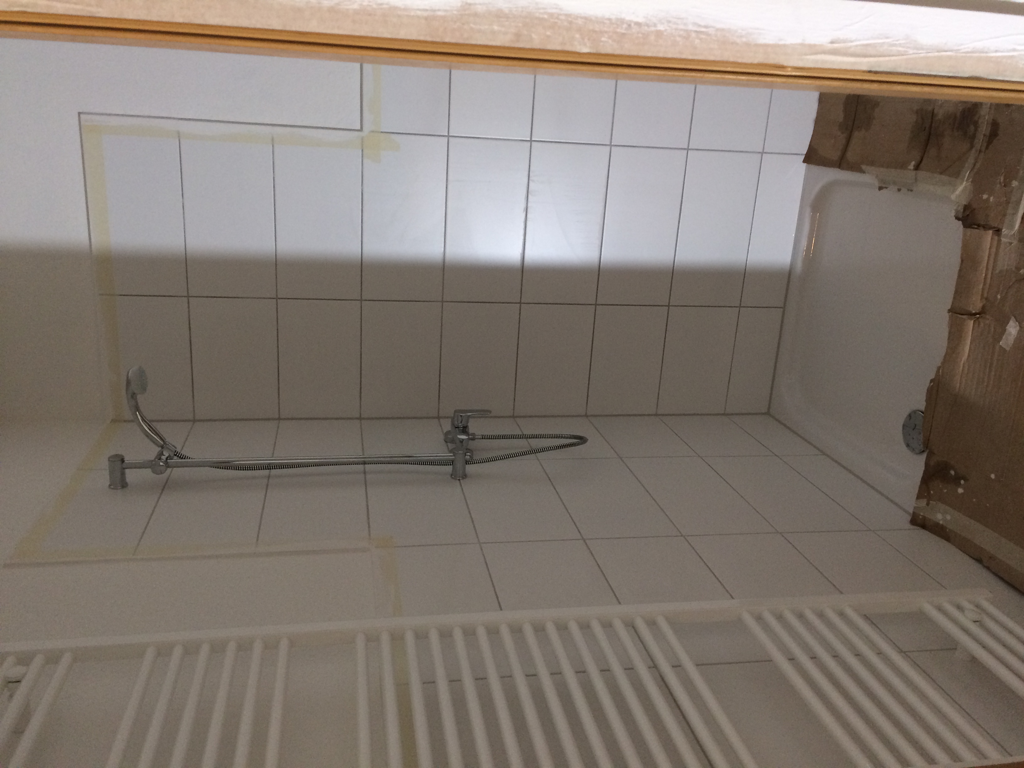 Białe płytki w łazience w trakcie remontu, widoczna bateria prysznicowa, grzejnik drabinkowy i zabezpieczona folią wanna.