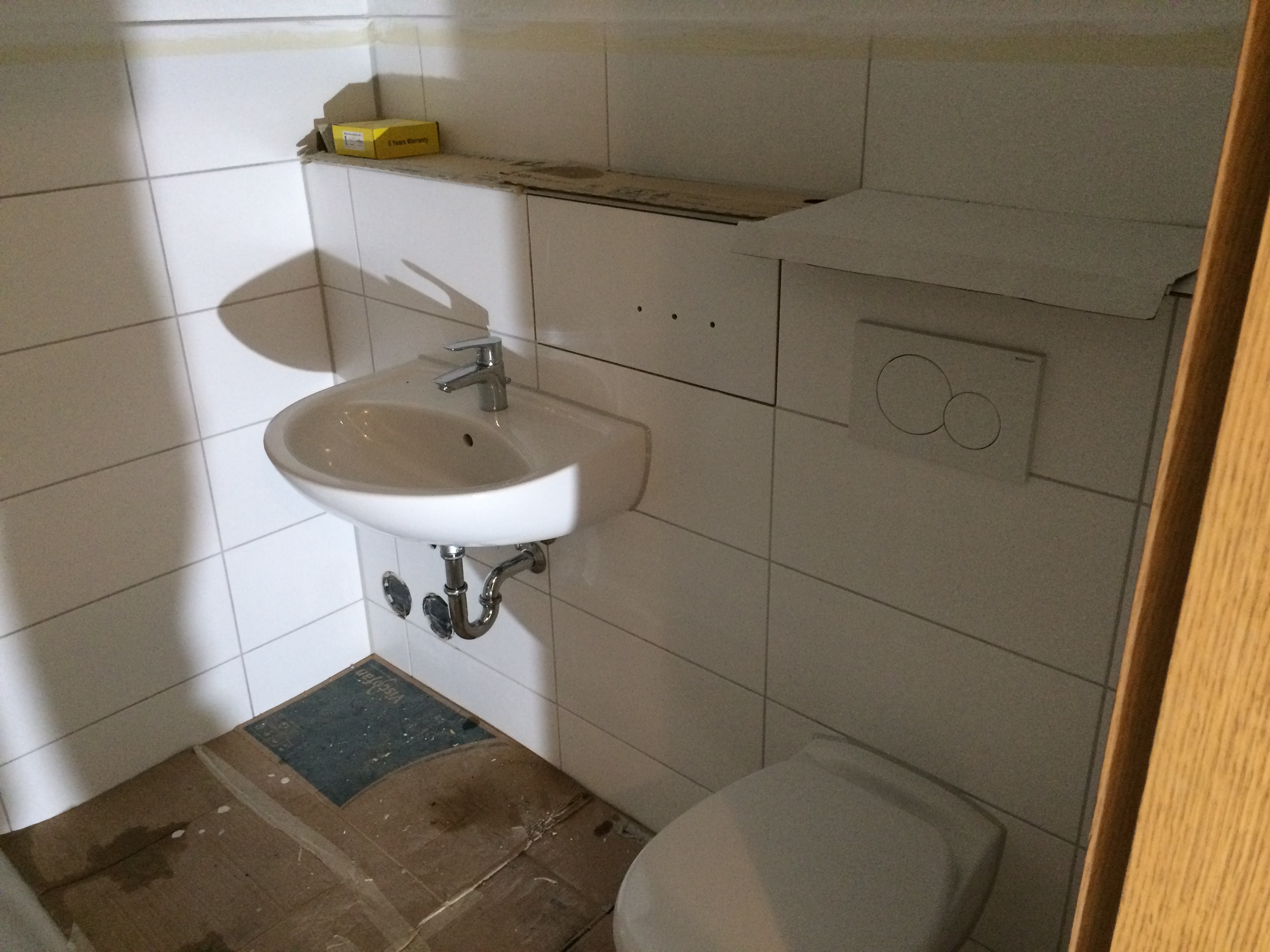 Biała umywalka zamontowana na ścianie z widocznymi rurami odpływowymi, biała toaleta, białe płytki na ścianach, karton na podłodze i żółte pudełko na półce w trakcie remontu łazienki.