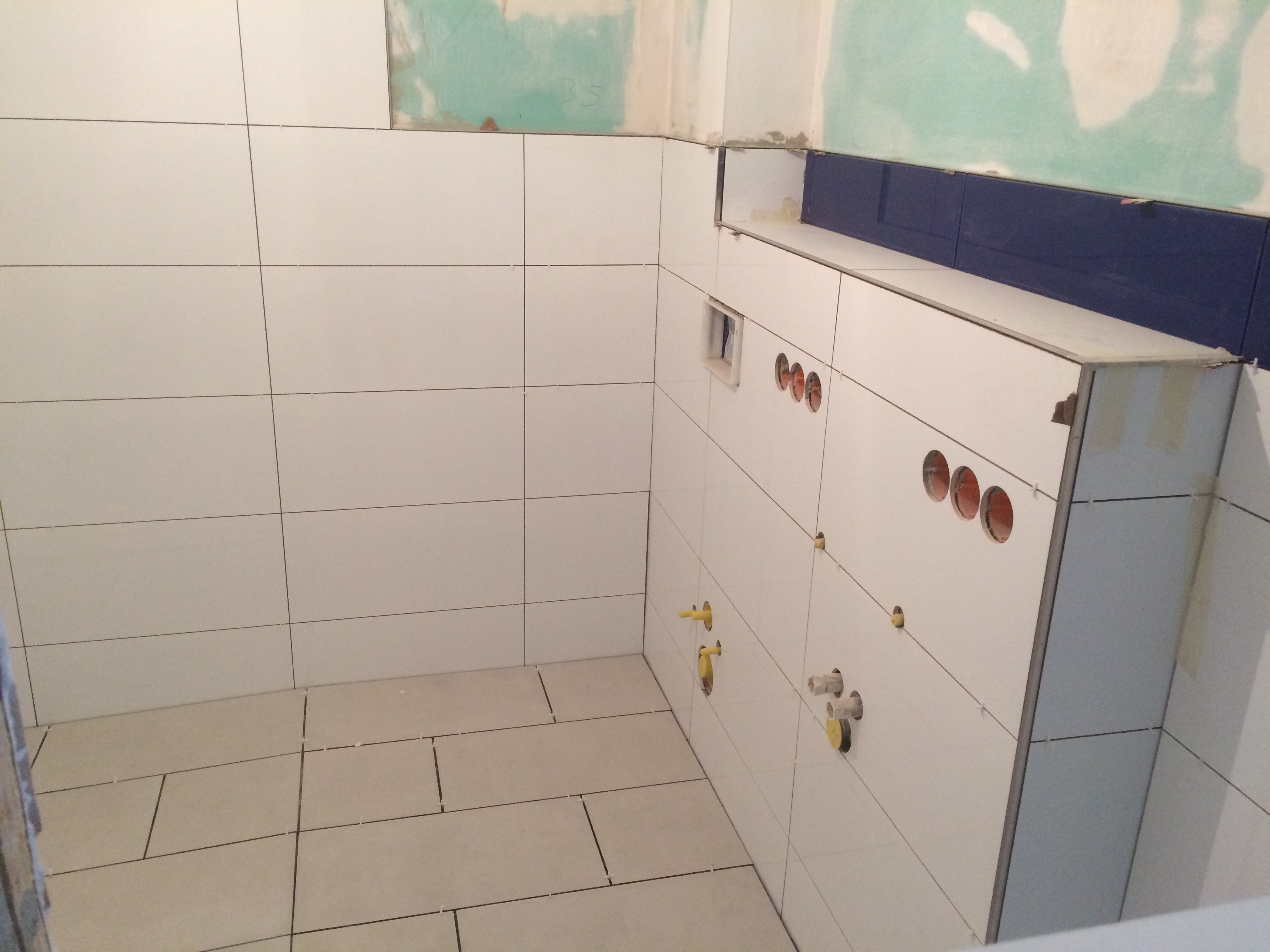 Białe płytki ceramiczne na ścianach i podłodze w łazience w trakcie montażu, widoczne otwory na rury i niebieski pas płytek na górze ściany.