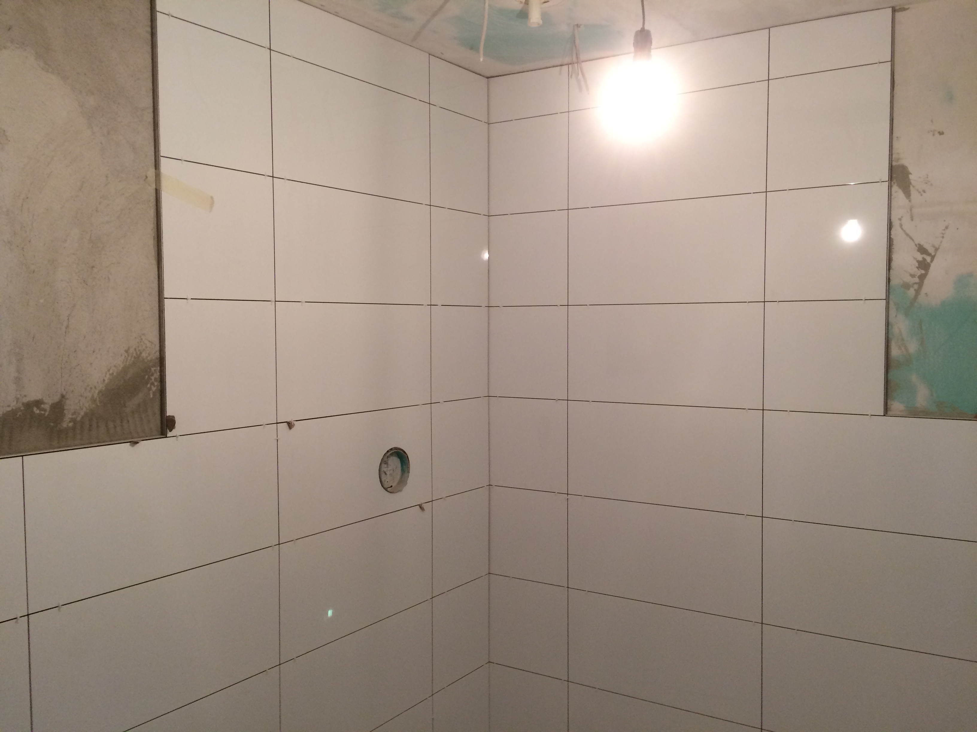 Ściana w trakcie układania białych, poziomych płytek z czarnymi fugami; widoczne niedokończone fragmenty ścian i otwór na instalację.