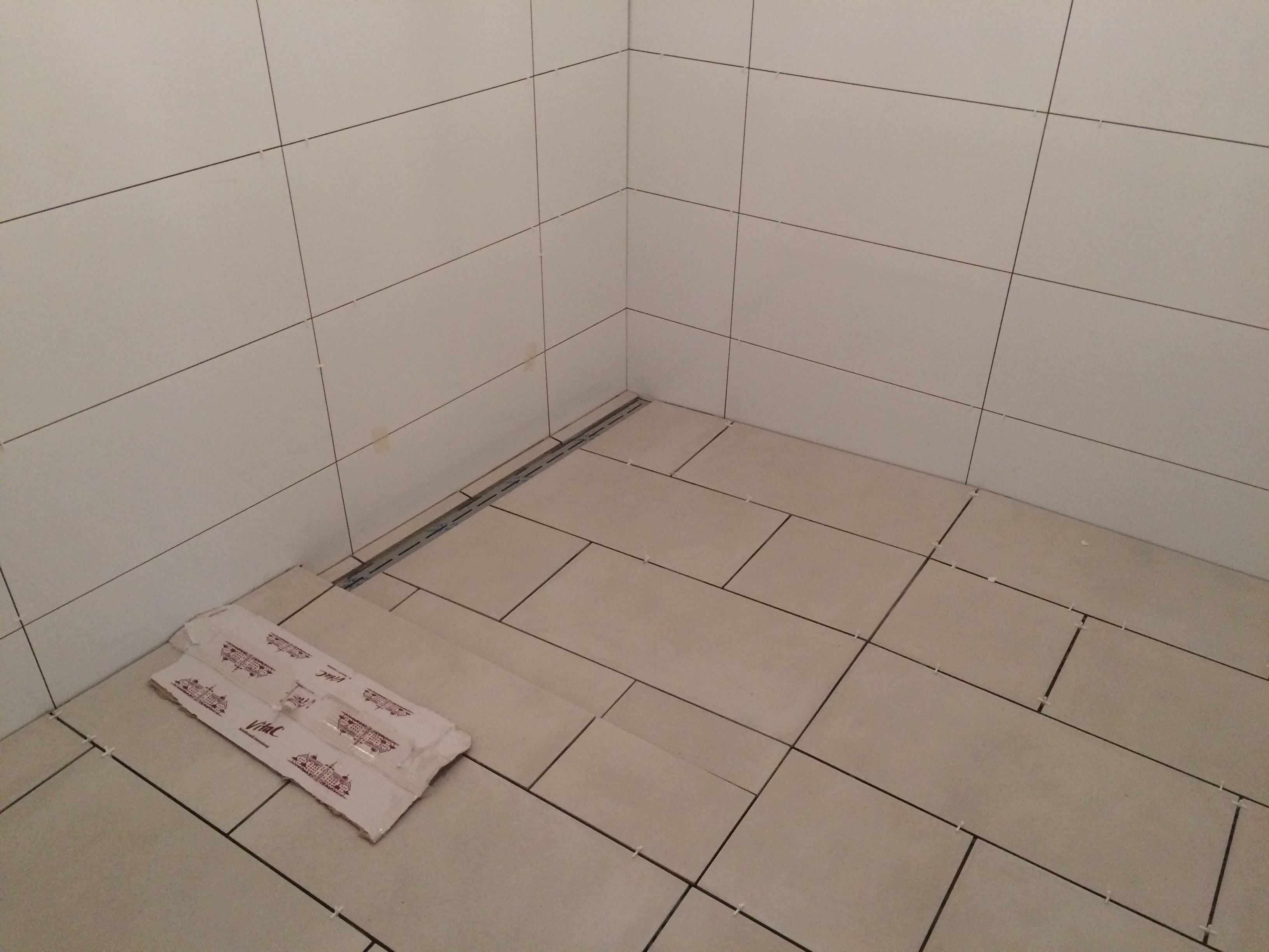 Wnętrze łazienki w trakcie układania beżowych płytek podłogowych i ściennych, widoczny odpływ liniowy i opakowanie krzyżyków dystansowych.