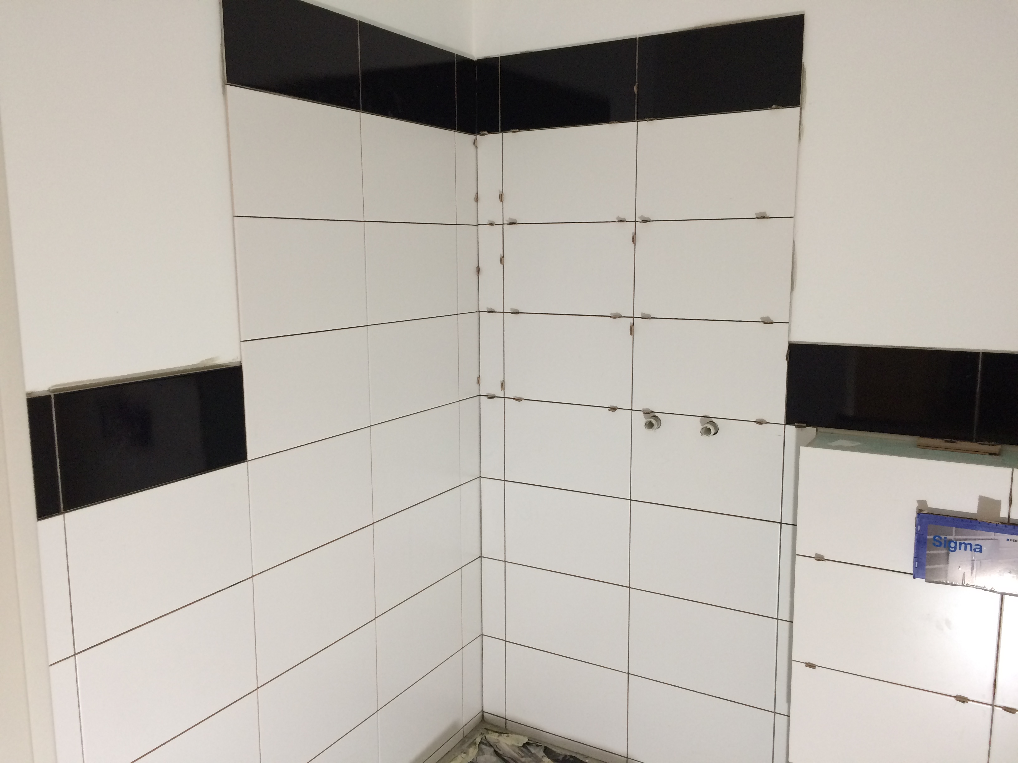 Ściana łazienki w trakcie wykańczania, z białymi płytkami i czarnym pasem u góry, widoczne krzyżyki dystansowe, przygotowane otwory na armaturę, dolny róg zabezpieczony folią.