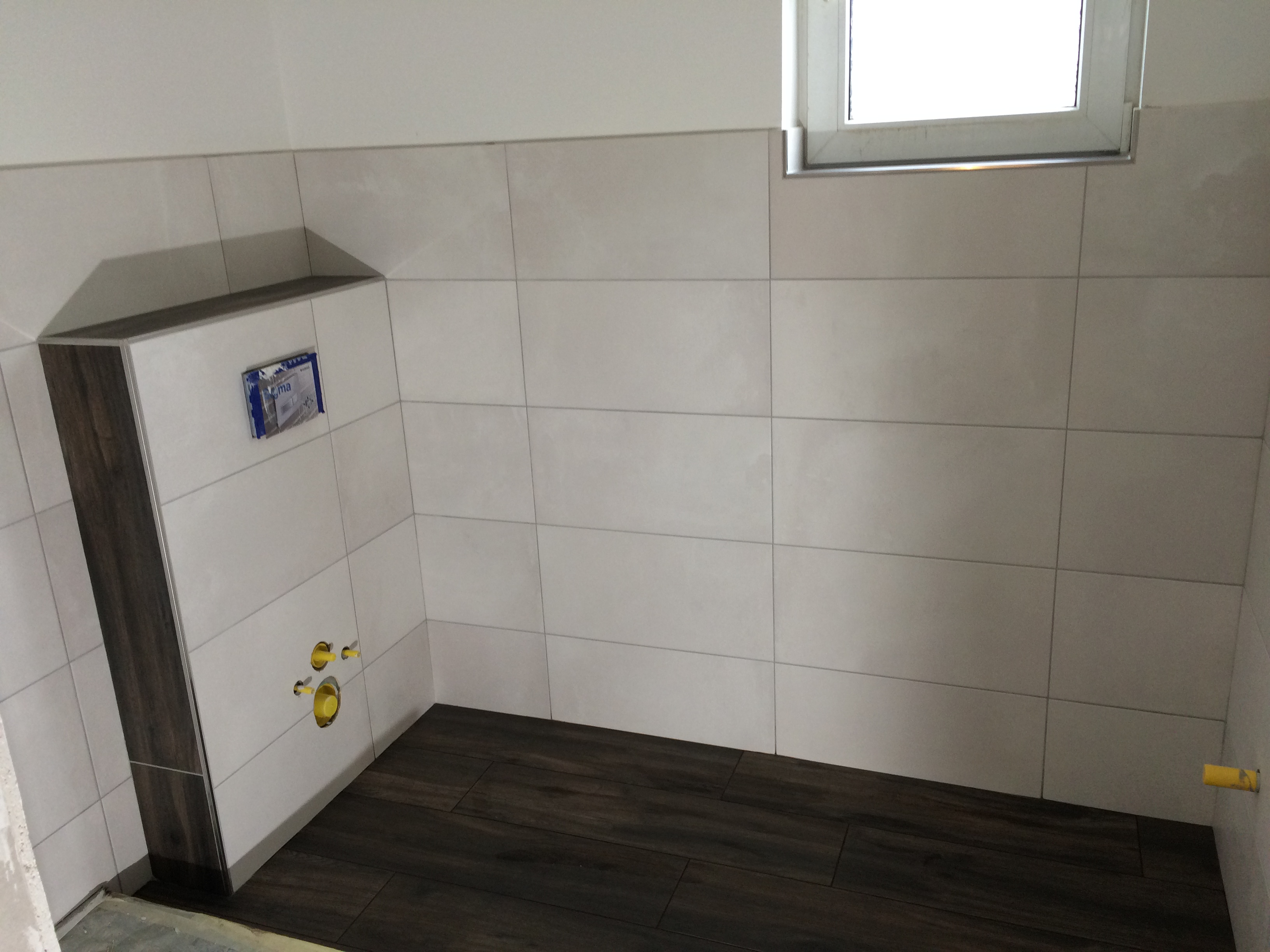 Wnętrze łazienki w trakcie wykańczania: ułożone jasne płytki ścienne i ciemne płytki podłogowe imitujące drewno, widoczne przyłącza wodne i stelaż podtynkowy.