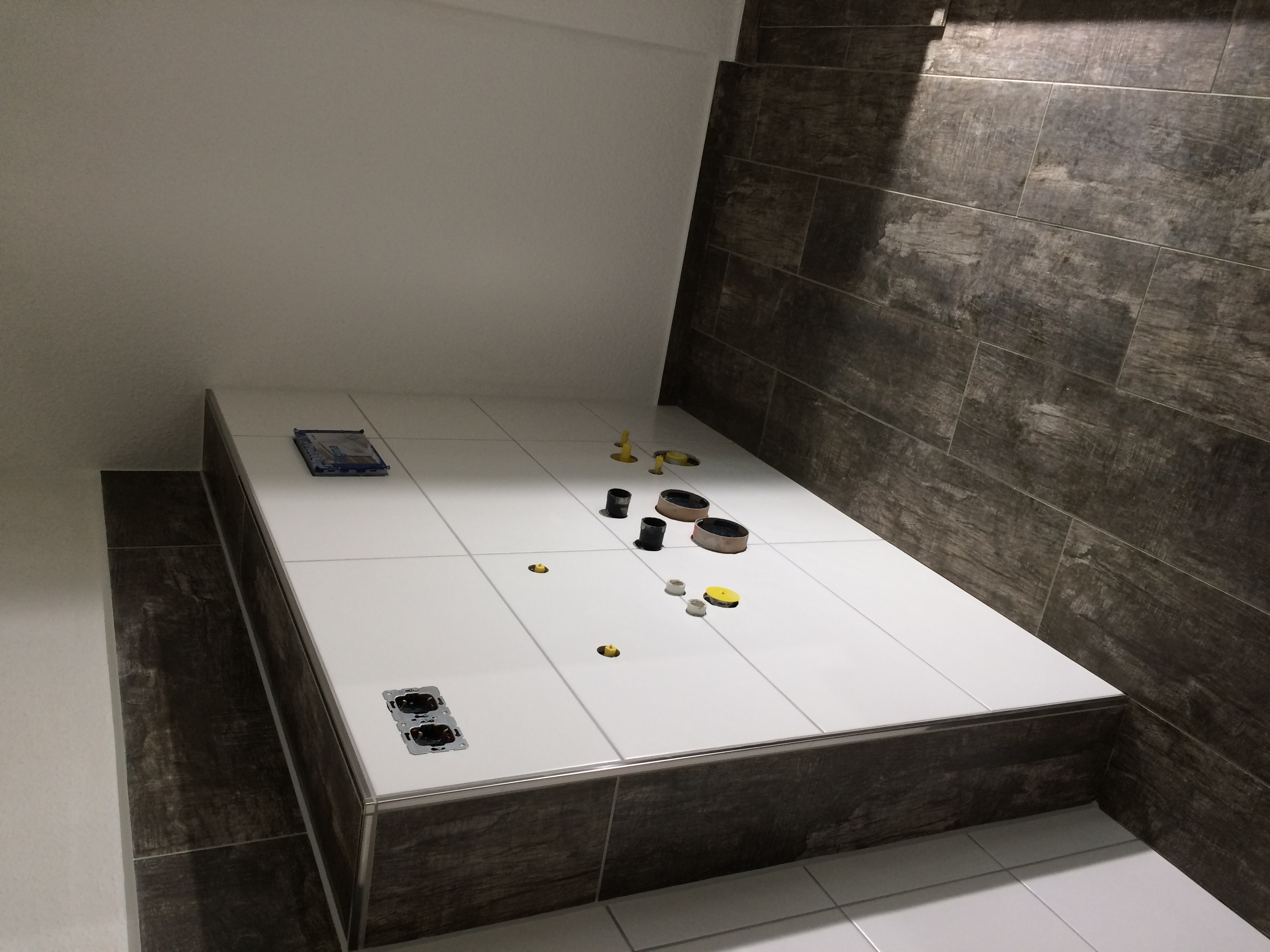 Podwyższony brodzik z białych płytek w trakcie montażu, widoczne otwory na odpływ i gniazdka elektryczne, ciemnobrązowe płytki na ścianie i obudowie brodzika.