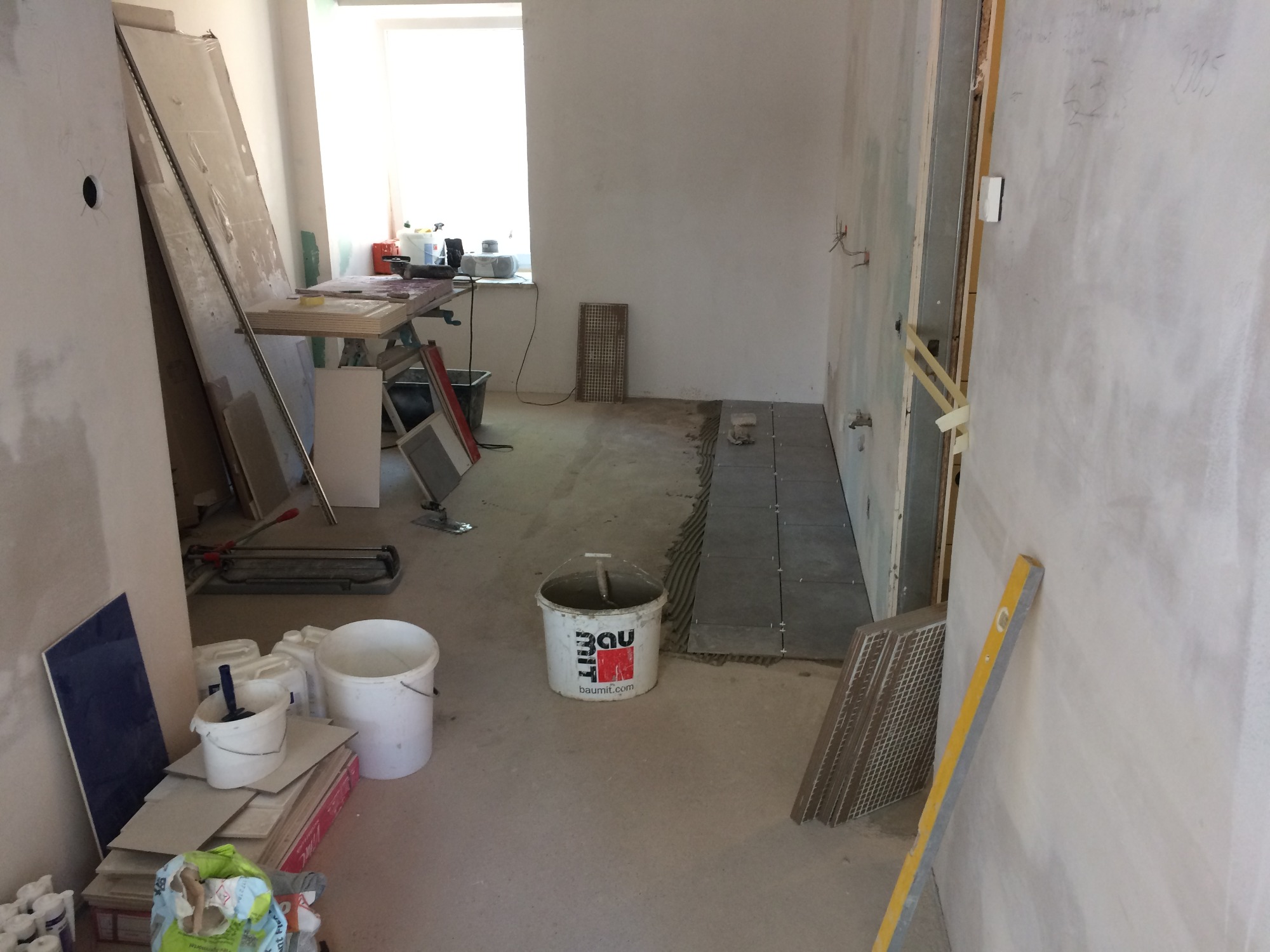 Wnętrze w trakcie układania szarych płytek podłogowych, widoczny kubeł z zaprawą Baumit, poziomica, narzędzia glazurnicze i przygotowane płyty gipsowe.