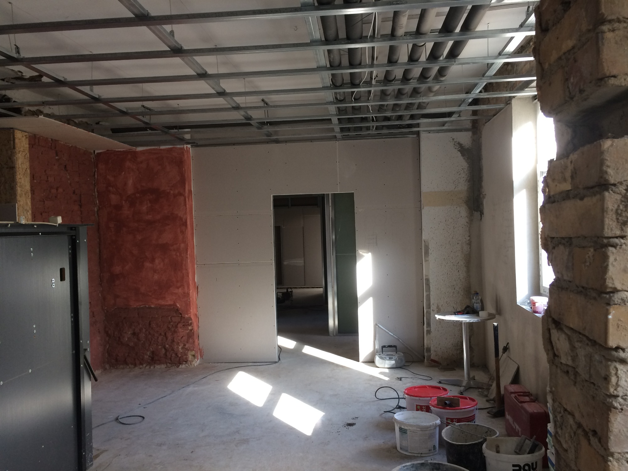 Wnętrze w trakcie prac wykończeniowych: widoczne profile sufitowe, płyty gipsowo-kartonowe na ścianach, fragmenty starej cegły i przygotowane wiadra z farbą na podłodze.