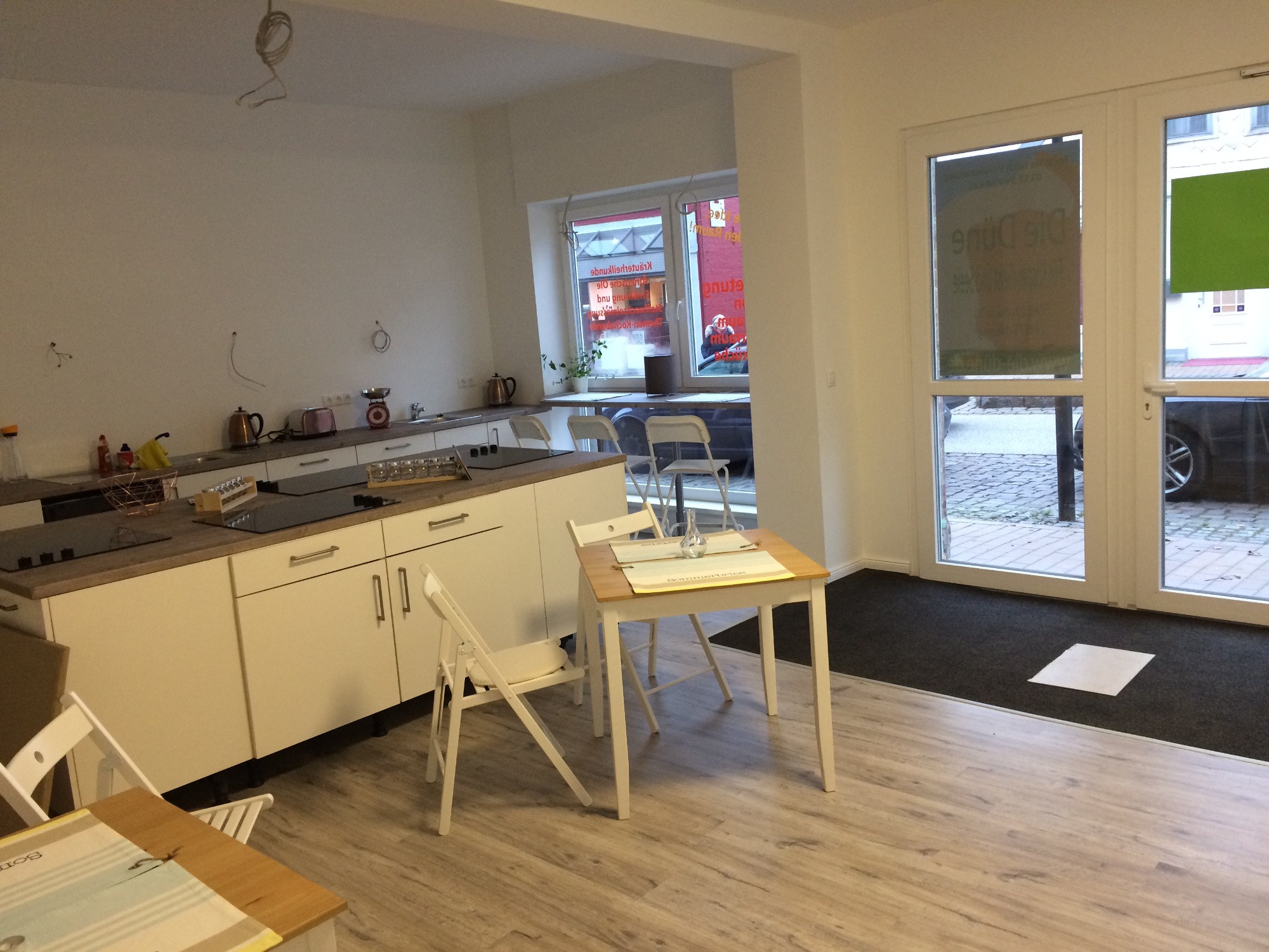 Jasna kuchnia w trakcie wykańczania, widoczne białe szafki z szarym blatem, stół z krzesłami, brakujące lampy i gniazdka na ścianach, okno z widokiem na ulicę.