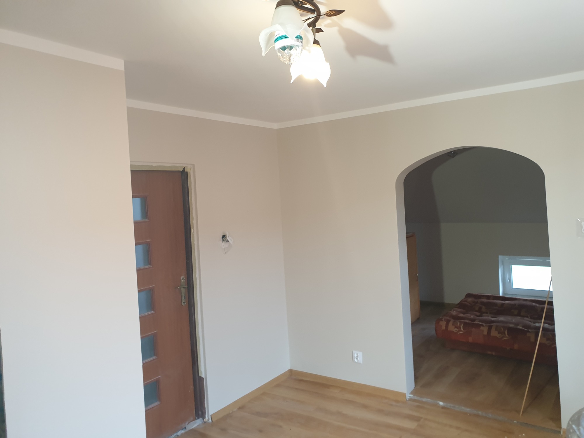 Wnętrze po malowaniu ścian na neutralny beż, widoczna drewniana podłoga, drzwi wejściowe z szybkami i łukowe przejście do pokoju z widocznym materacem i oknem.