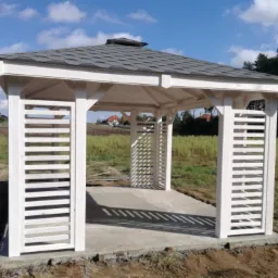 Biała, drewniana altana z szarym, dachówkowym dachem, widoczna na tle zielonego pola i niebieskiego nieba, z elementami zabudowy w oddali.