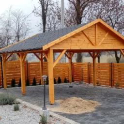 Drewniana pergola z szarym dachem z gontu bitumicznego, widoczna z zewnątrz, otoczona drewnianym płotem i kostką brukową, z małym kopcem piasku w środku.