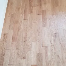 Ułożona podłoga z paneli imitujących drewno, widoczne łączenia i wzór słojów, listwa przypodłogowa przy ścianie, fragment grzejnika.