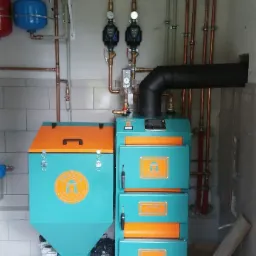 Turkusowo-pomarańczowy piec na paliwo stałe z podajnikiem i rurami miedzianymi w kotłowni z szarą podłogą.