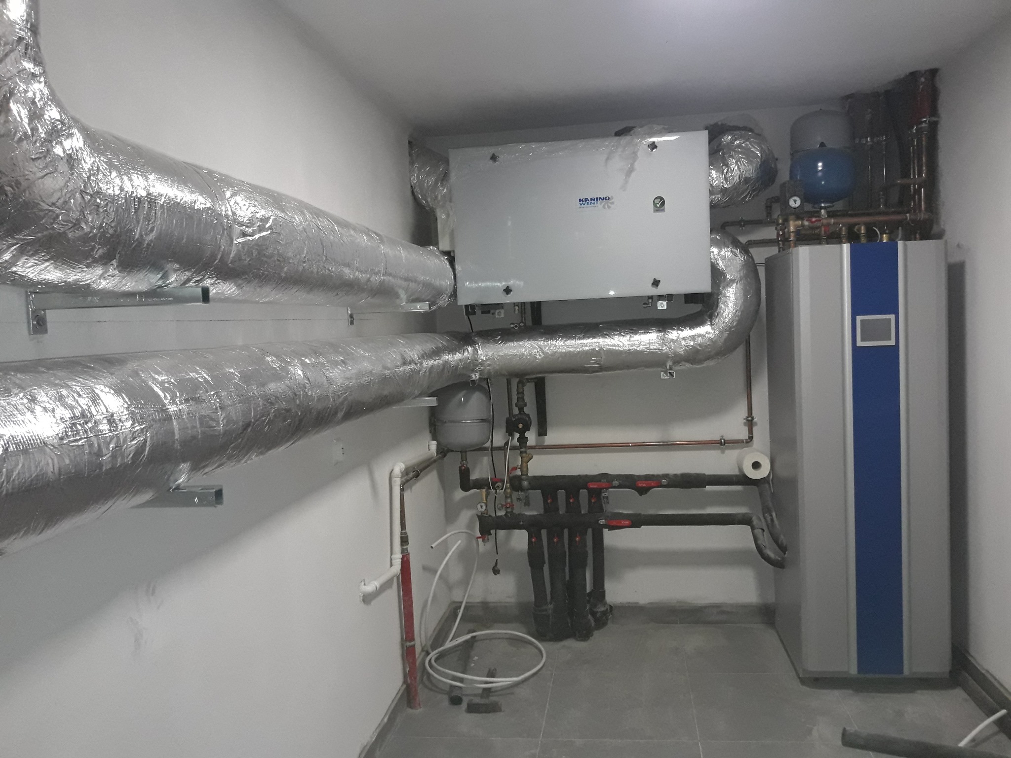 Instalacja wentylacyjna z rurami w izolacji aluminiowej, centrala wentylacyjna marki Karino West w folii ochronnej, oraz pion instalacji grzewczej z czarnymi zaworami i zbiornikiem wyrównawczym...