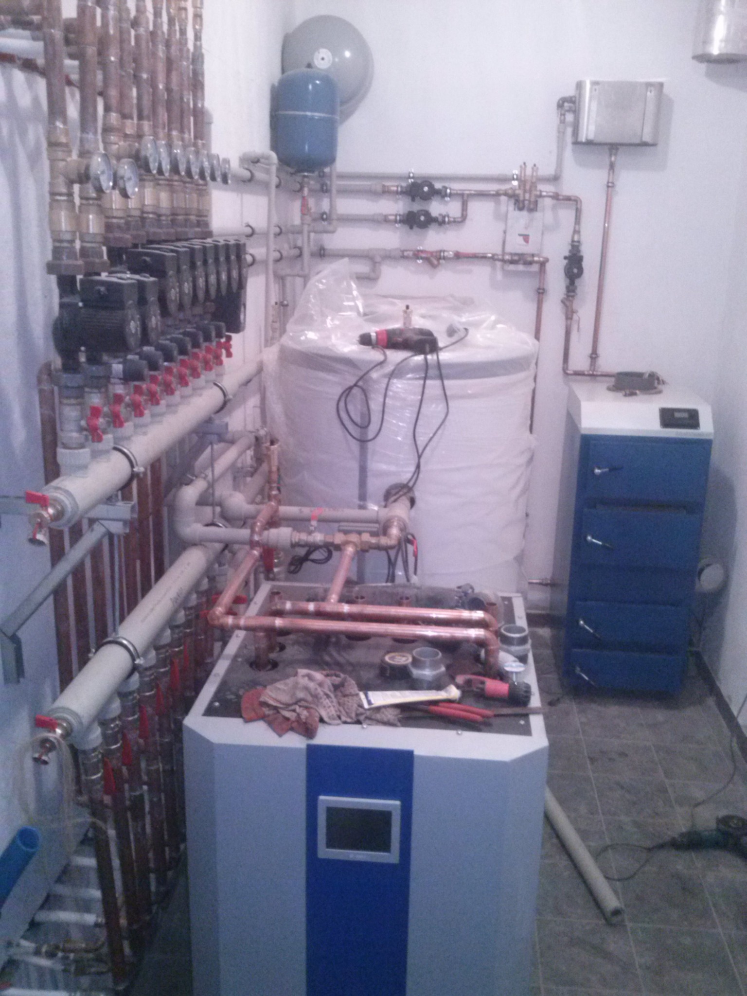 Węzeł cieplny w kotłowni, widoczne liczne rury miedziane, zawory, pompy obiegowe, zasobnik ciepłej wody owinięty folią, narzędzia do lutowania na górze kotła, oraz elektryczne narzędzie...