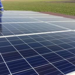 Solar-Volt s.c - Nowe panele słoneczne z widocznym fragmentem pracownika w niebieskim kombinezonie, zamontowane na brązowym dachu, z zielonym polem w tle.