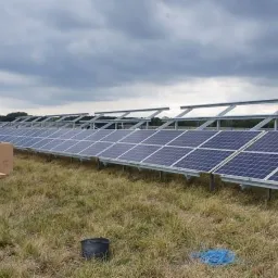 Montaż farmy fotowoltaicznej: rzędy paneli na metalowej konstrukcji, kartony z logo Q CELLS, zachmurzone niebo w tle.