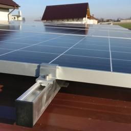 Solar-Volt s.c - Panele słoneczne zamontowane na brązowym dachu z widocznymi aluminiowymi szynami montażowymi i mocowaniami, z fragmentem sylwetki osoby w niebieskim kombinezonie w tle.