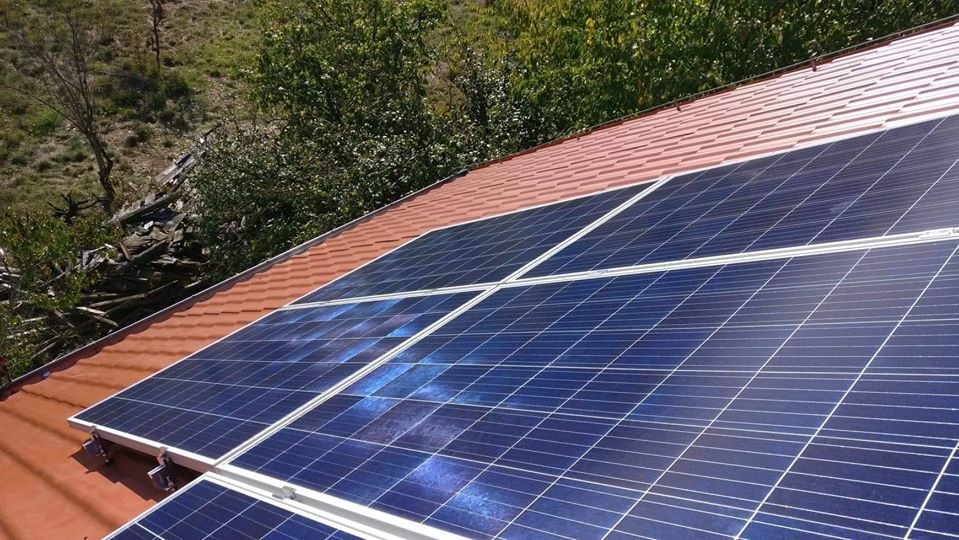 Panele słoneczne na dachu pokrytym czerwoną dachówką bitumiczną, widok z góry, w tle zieleń i fragmenty rozbiórki.