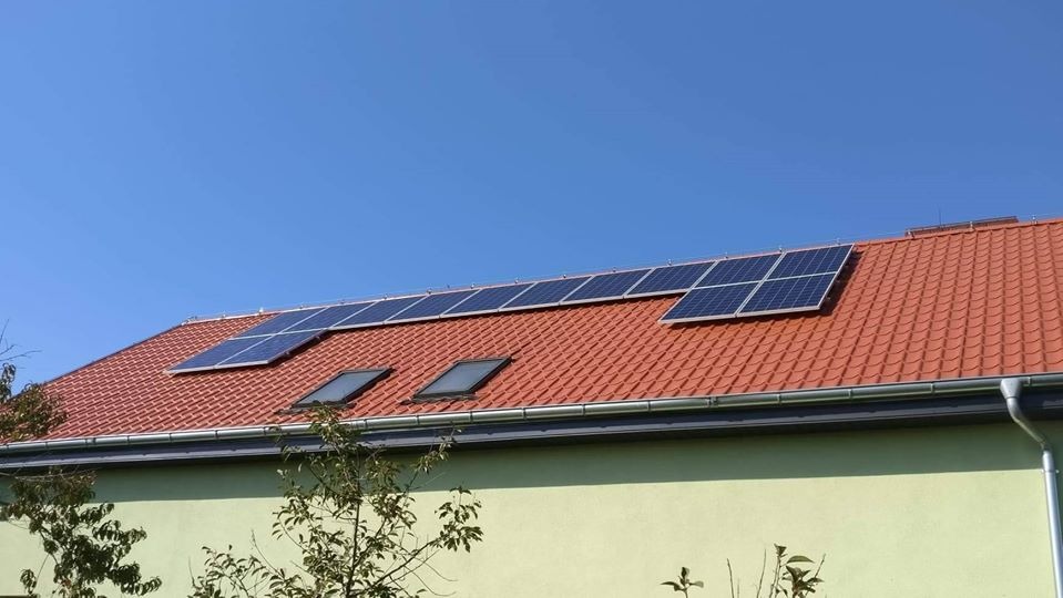 Czerwony dach z dachówką ceramiczną, na którym zamontowano panele słoneczne, oraz dwa okna dachowe, widok z dołu na tle błękitnego nieba.