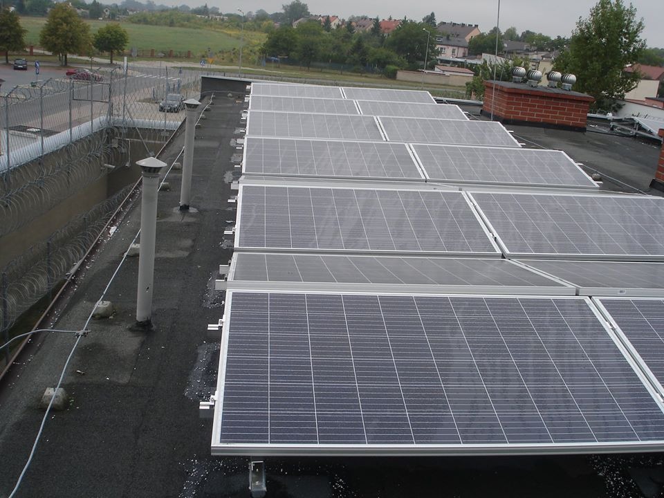 Rząd paneli słonecznych na płaskim dachu budynku z widocznym ogrodzeniem z drutu kolczastego i kominem w tle.