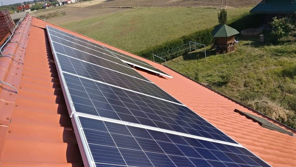 Panele słoneczne zamontowane na dachu z czerwonej dachówki, widok z boku, w tle zielony ogród z altaną.